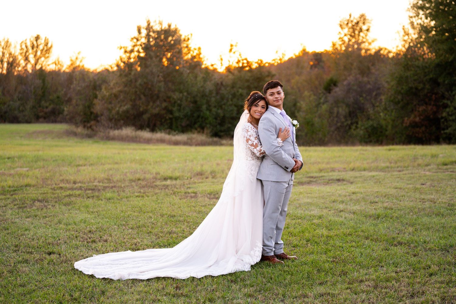 Wedding couple poses in field during golden hour with flowing white dress train.