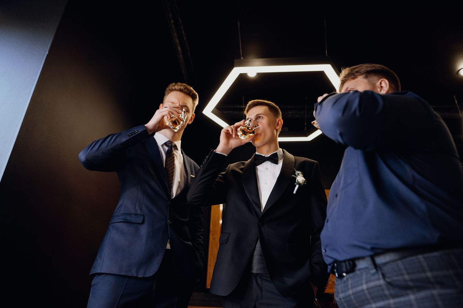 Groomsmen in dark suits share a celebratory moment while getting ready for a wedding in a dimly lit room.