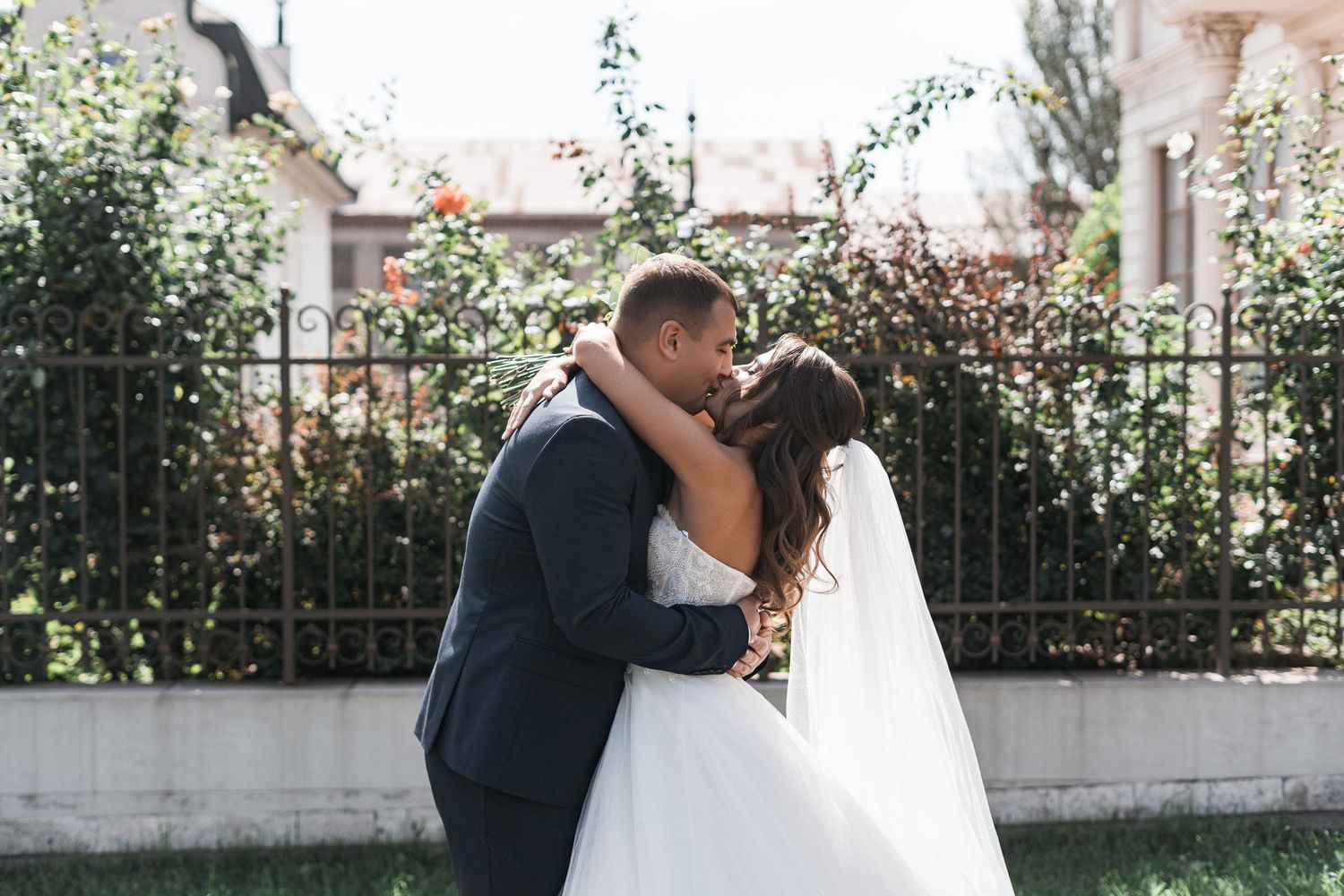 A romantic couple in wedding attire embraces in a sunlit garden with a white stone wall and greenery backdrop.