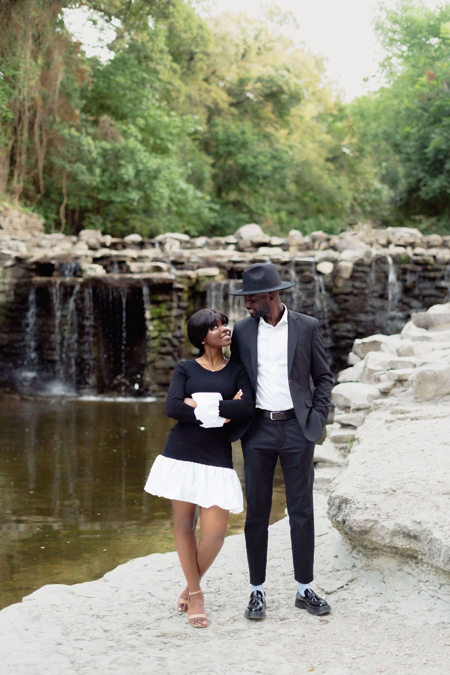 A stylishly dressed couple walks along a rocky waterfall path in coordinated black and white outfits.