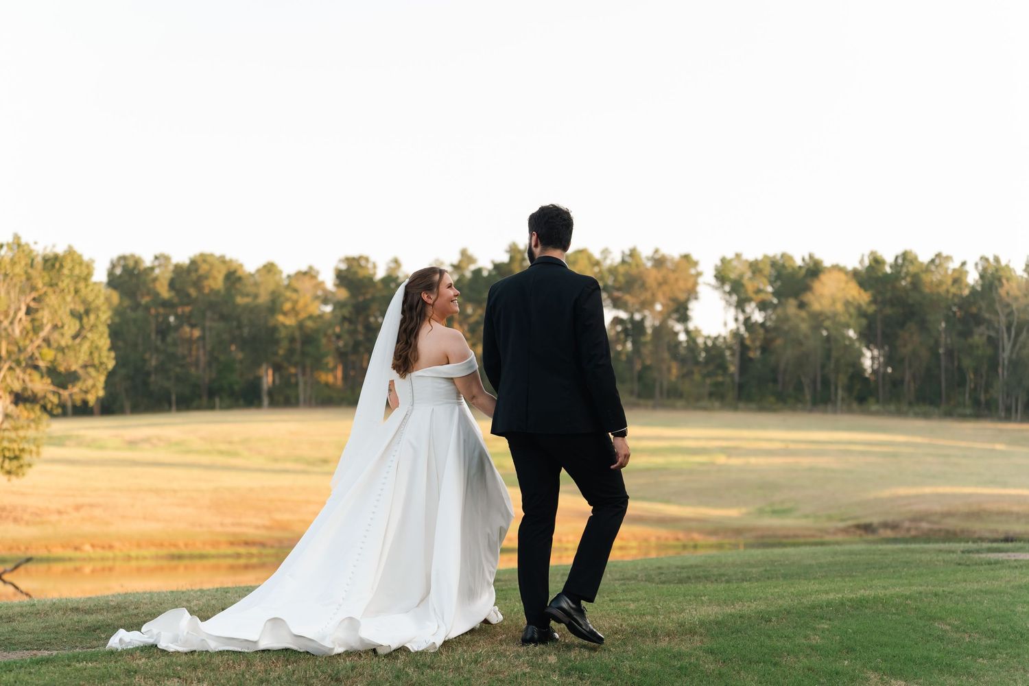 A couple in wedding attire stands together on a scenic golf course at sunset.