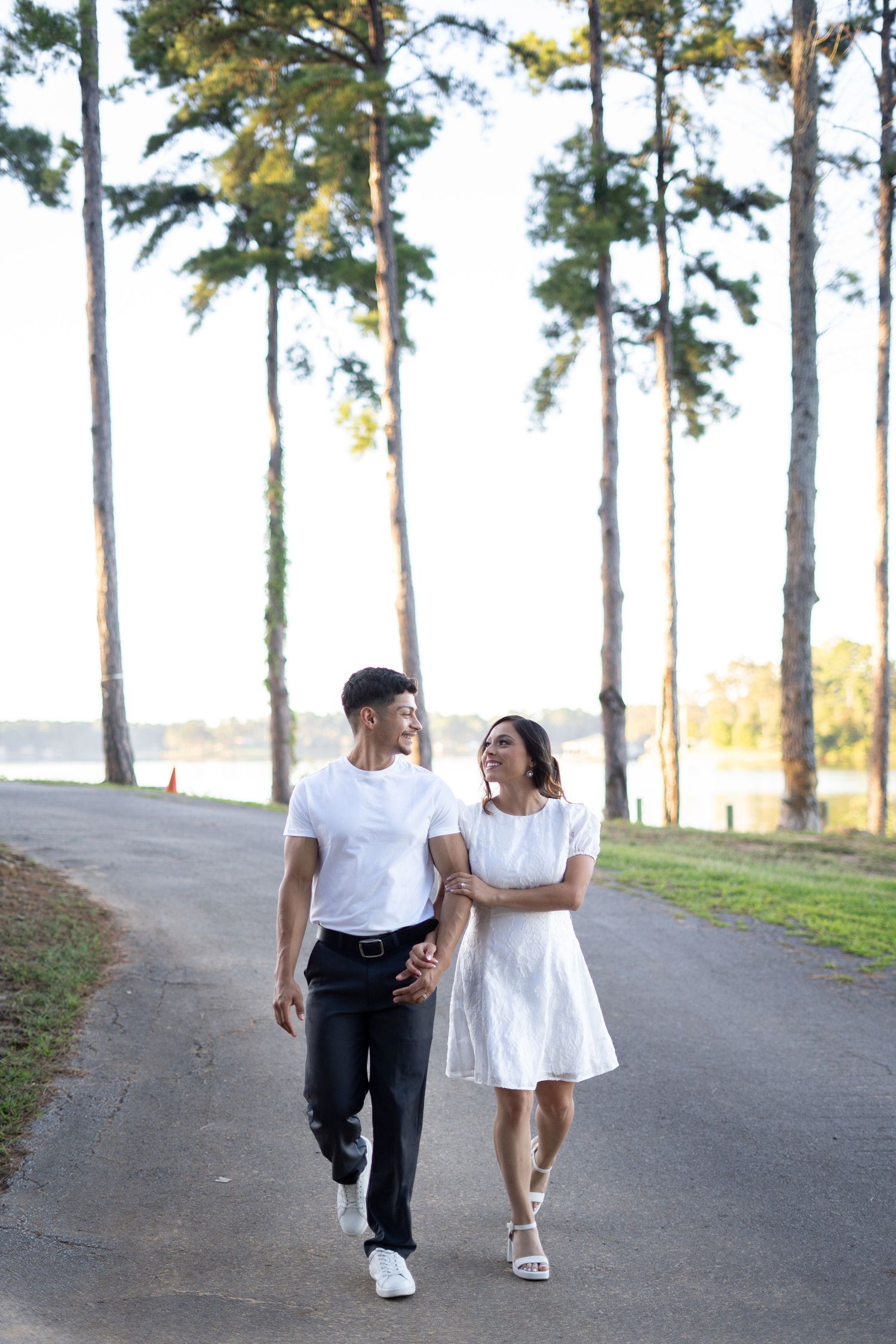 Couple in white clothing walks and laughs together on tree-lined path.