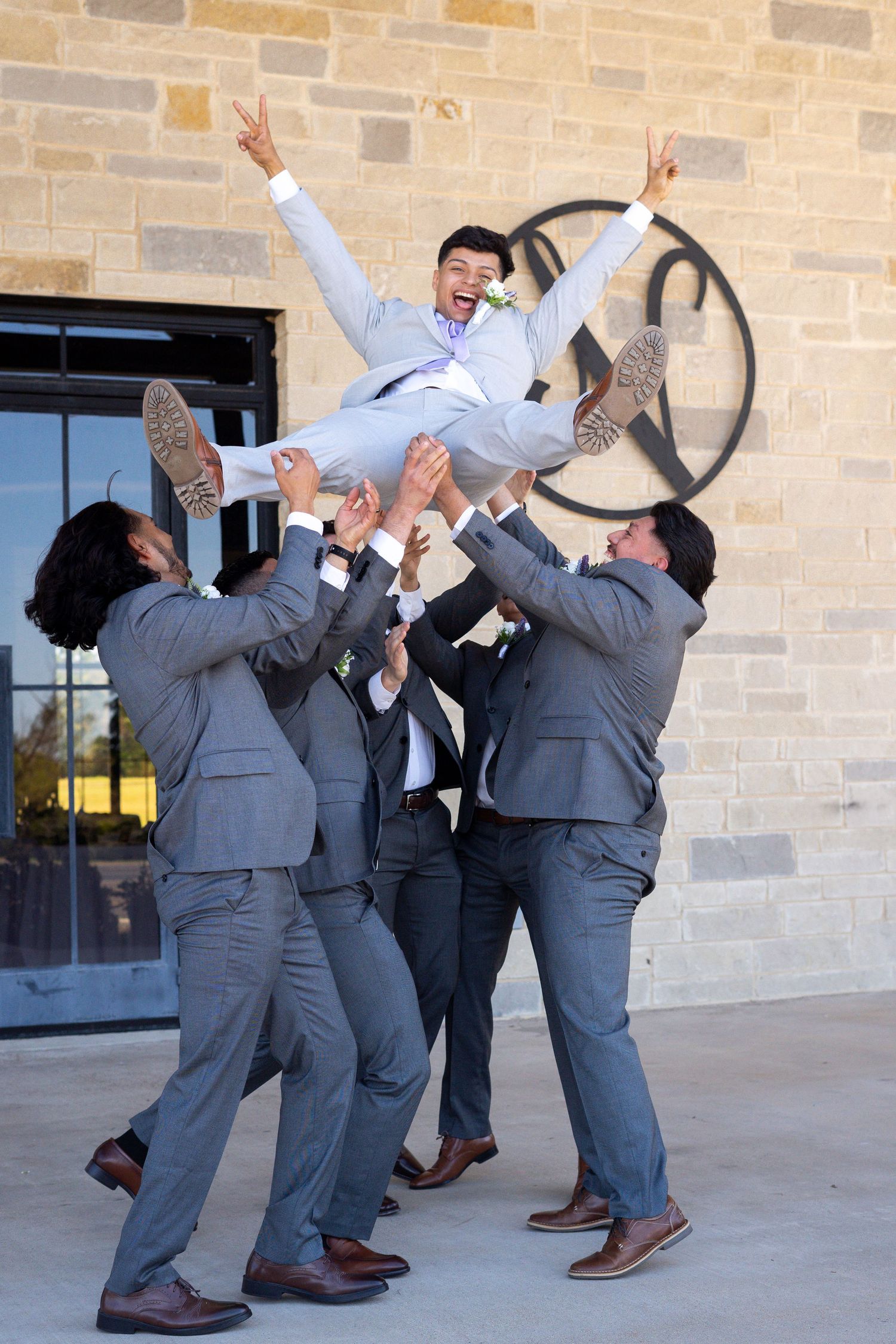 Groomsmen in gray suits perform playful jumping poses against a brick wall during wedding photos.