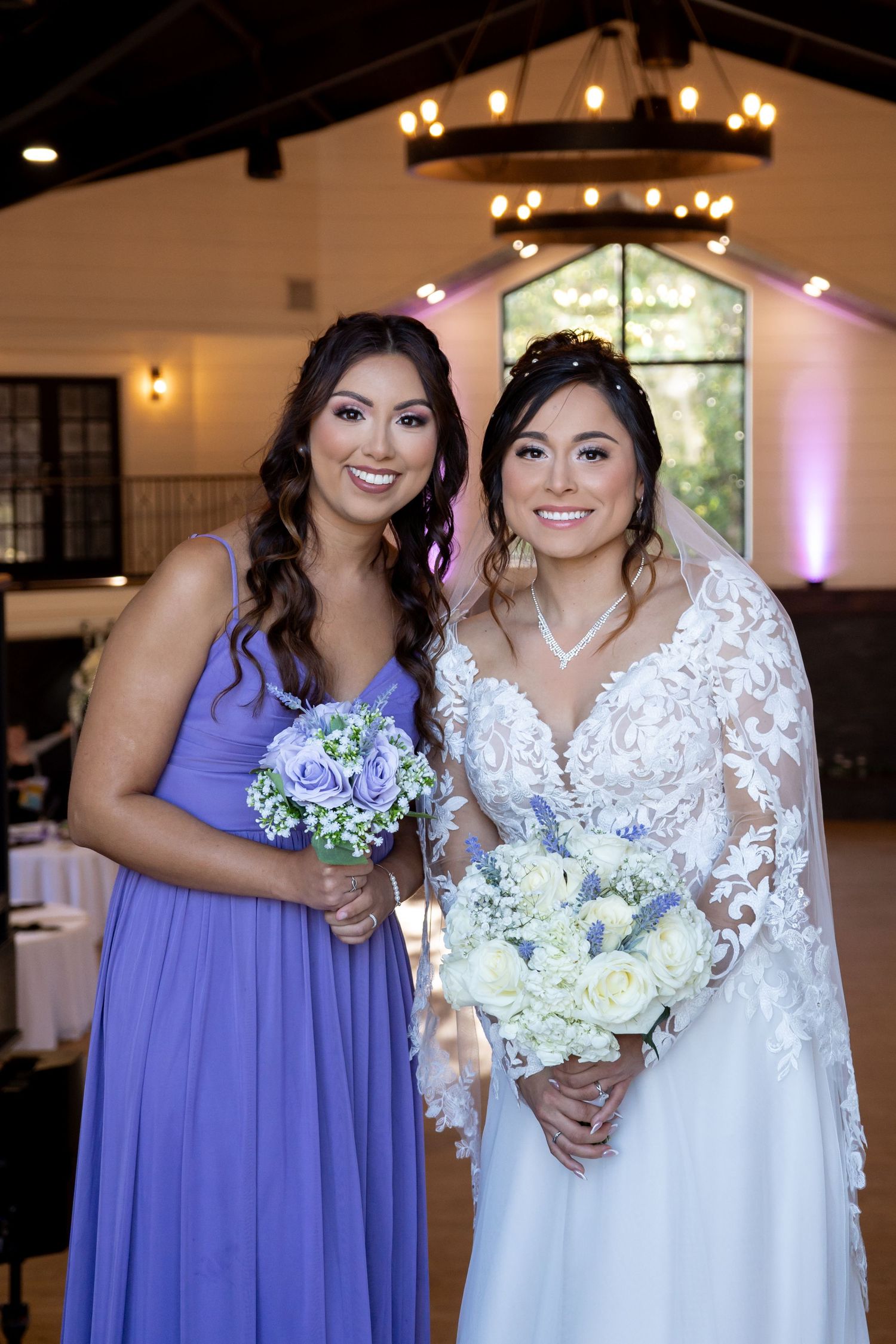 Wedding party members in white and lavender dresses smile while holding bouquets in a reception venue.