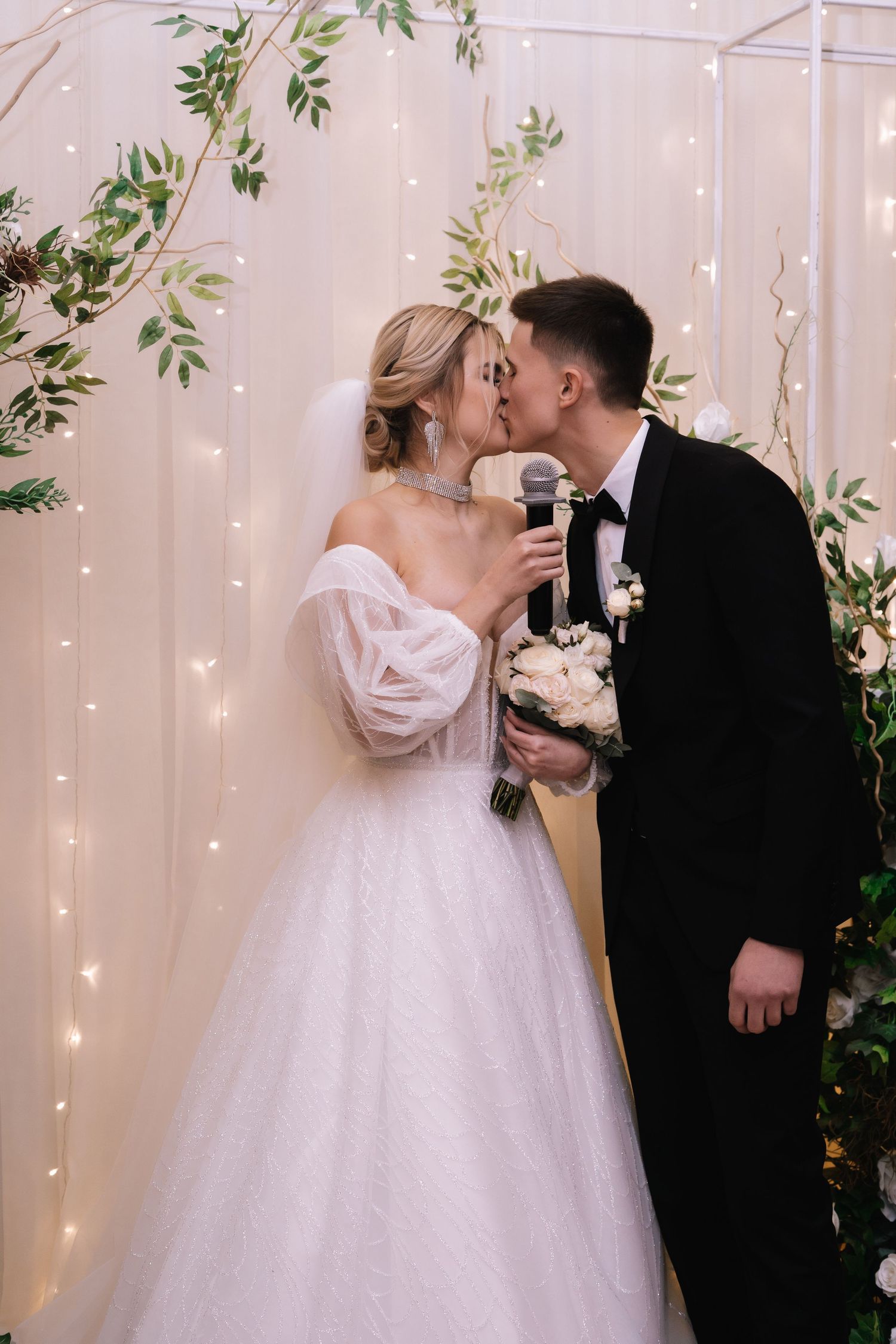 A bride and groom share a romantic kiss in front of a fairy light backdrop with greenery decorations at their wedding.
