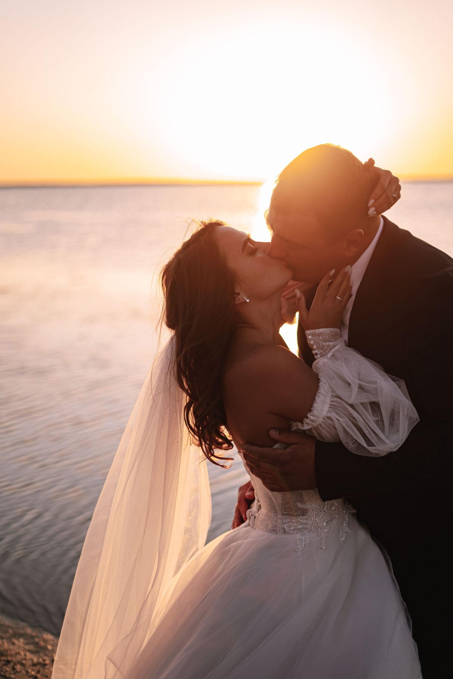 A romantic silhouette of newlyweds embracing against a golden sunset over calm waters with a flowing bridal veil.