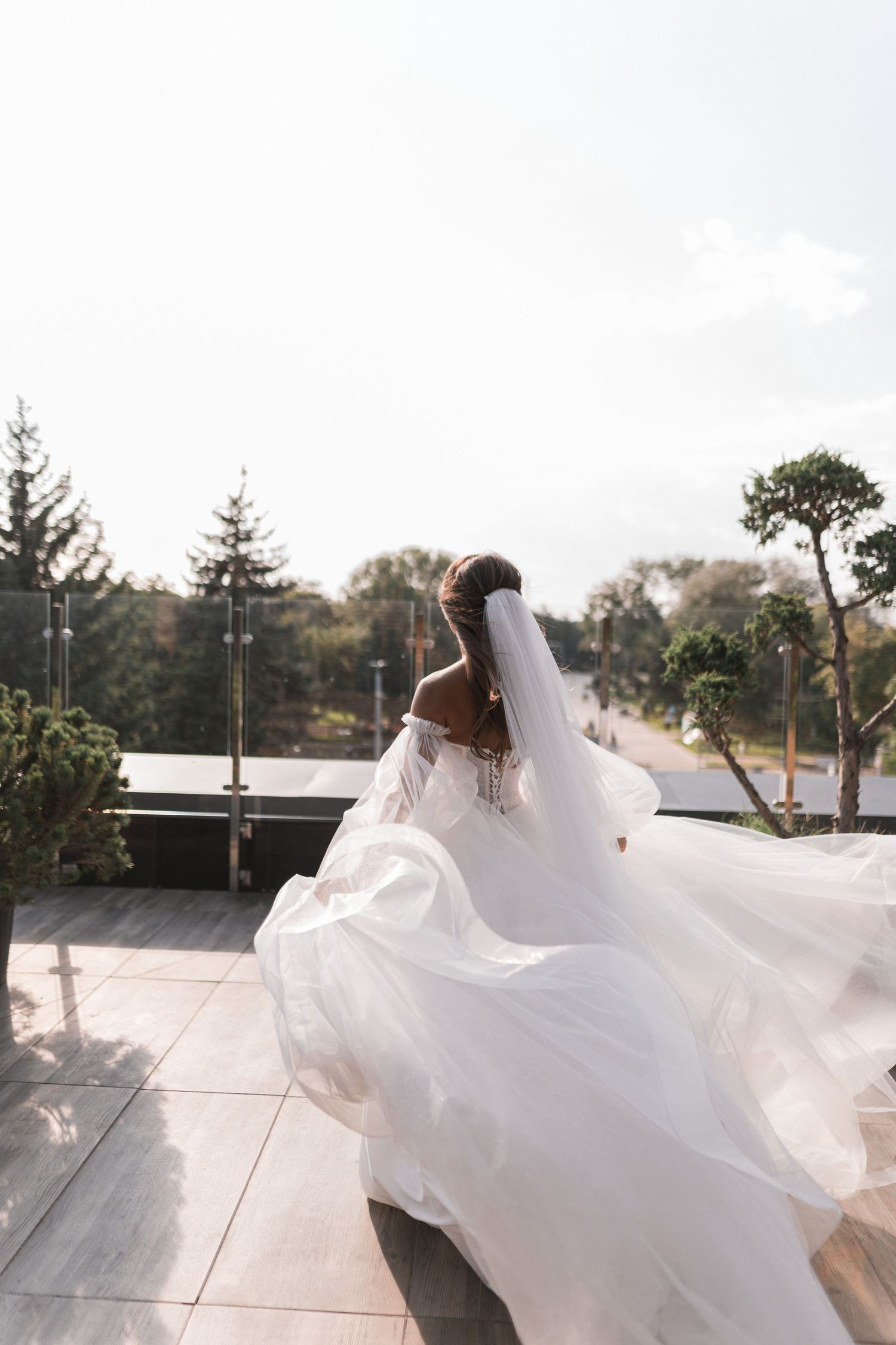 A bride in a flowing white dress twirls on a rooftop terrace creating a dramatic movement with her veil in the sunlight.