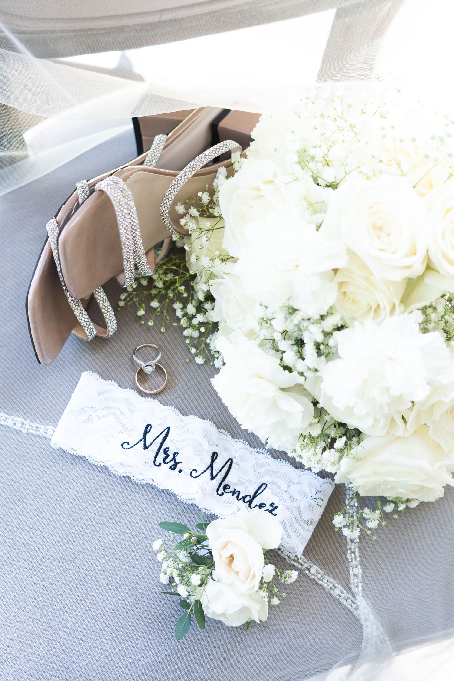 Wedding details including sparkly shoes, wedding rings, name card and white rose bouquet arranged on gray background.