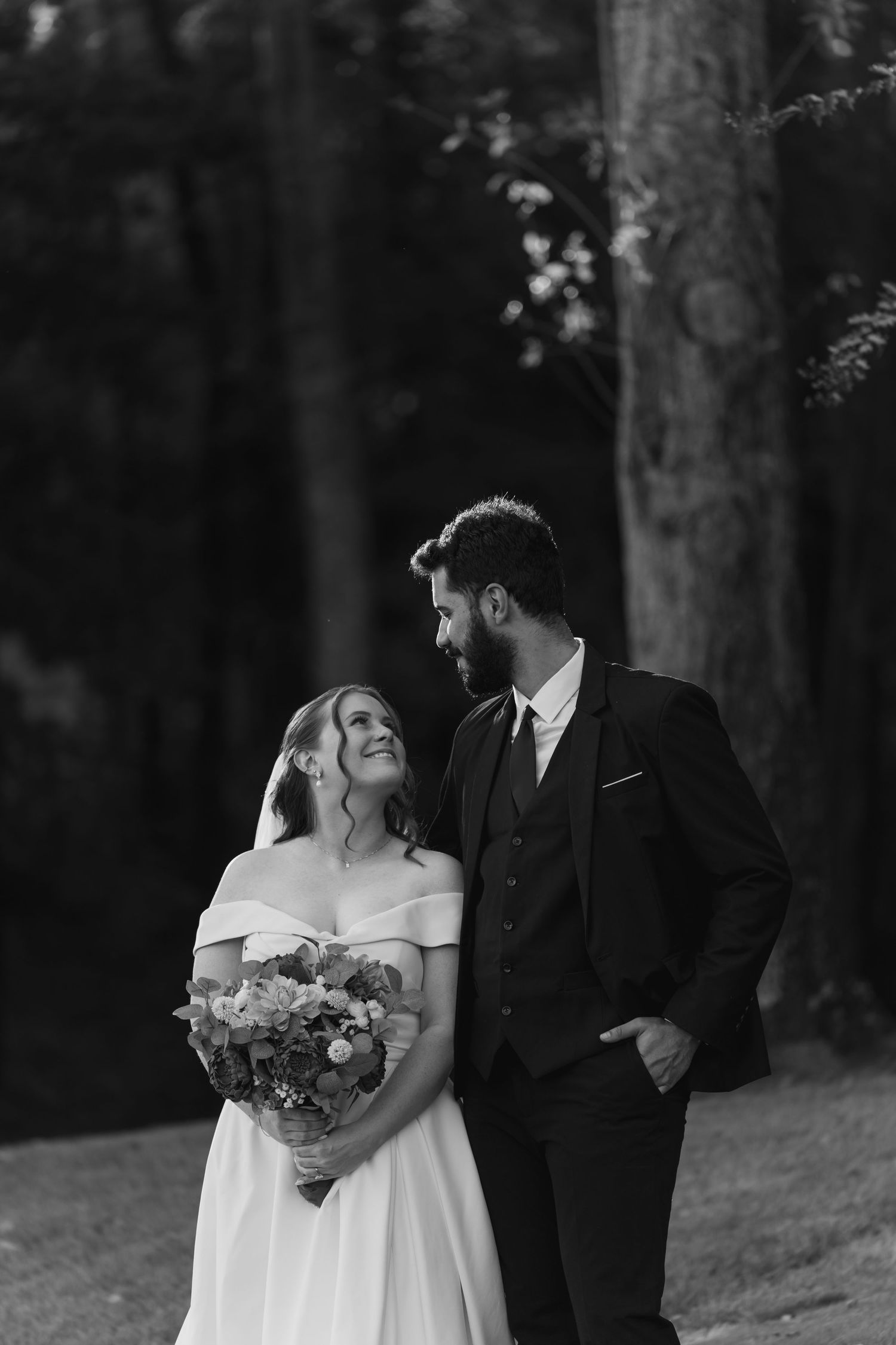 A romantic black and white wedding portrait shows a couple in formal attire standing among trees.