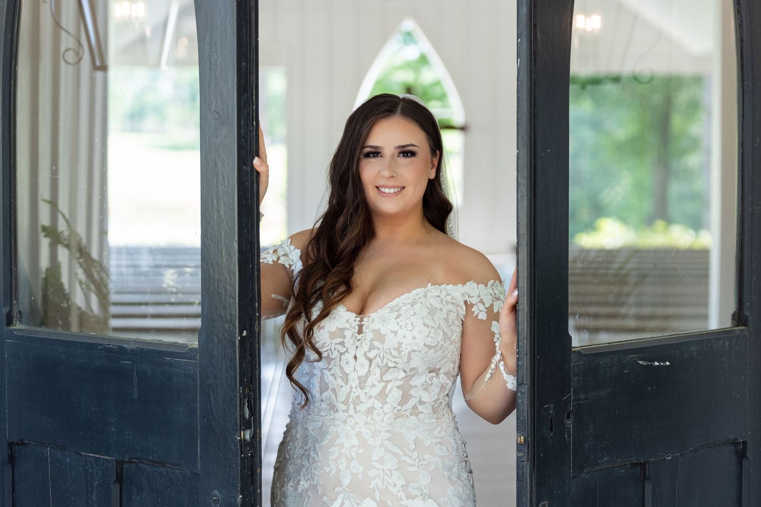 A bride in a beaded off-shoulder wedding gown stands in a doorway with a bright smile.