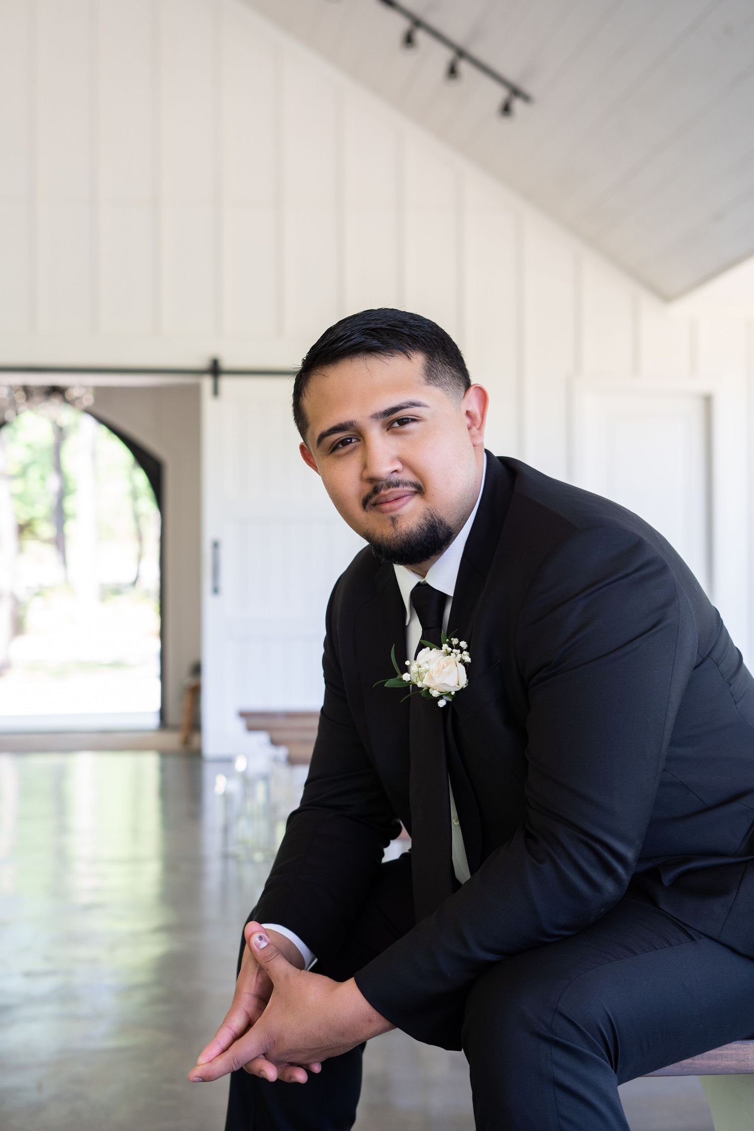 A person in a formal black suit with a white boutonniere sits posed in an elegant venue with arched doorways.