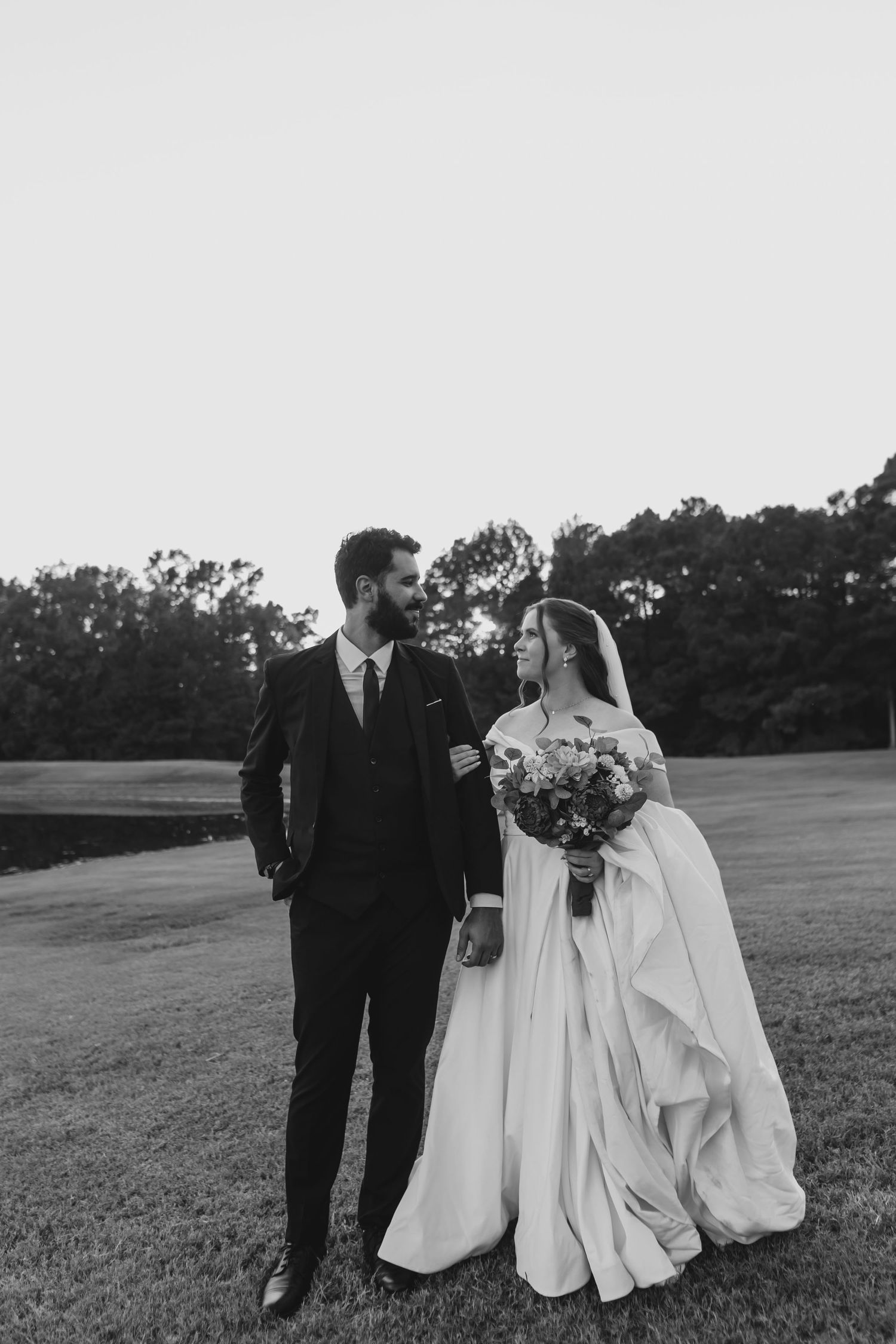 A black and white photo of a couple walking together on a grassy field with trees in the background.