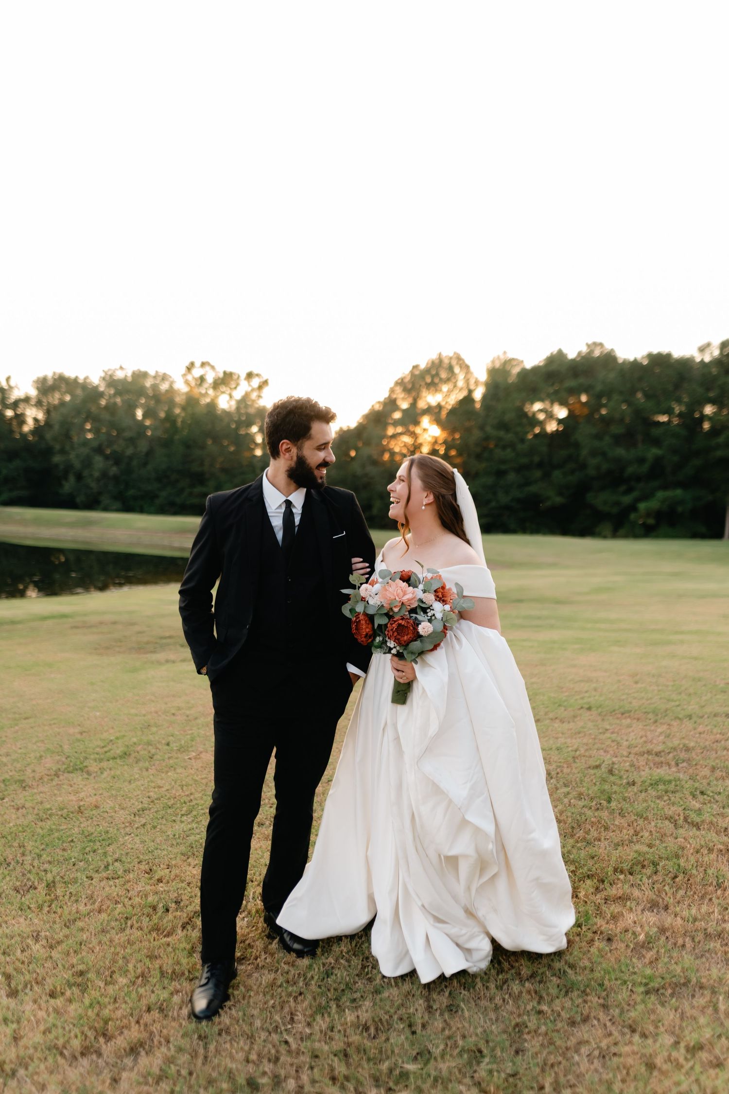 A romantic wedding couple walking through a golden field at sunset in formal attire.