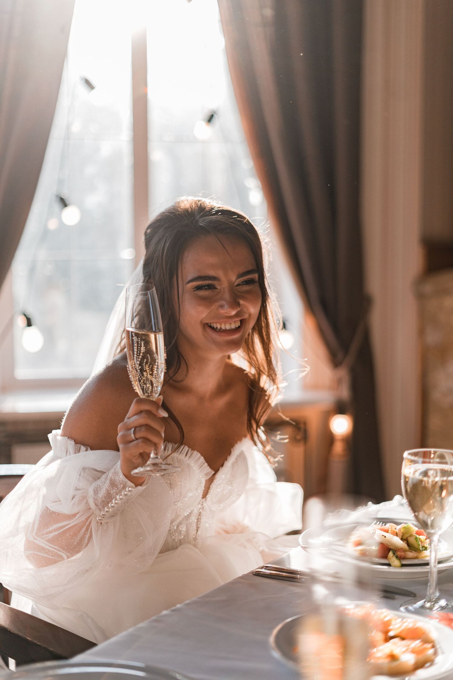 A bride in a white off-shoulder dress smiles while holding a champagne glass during a wedding reception.