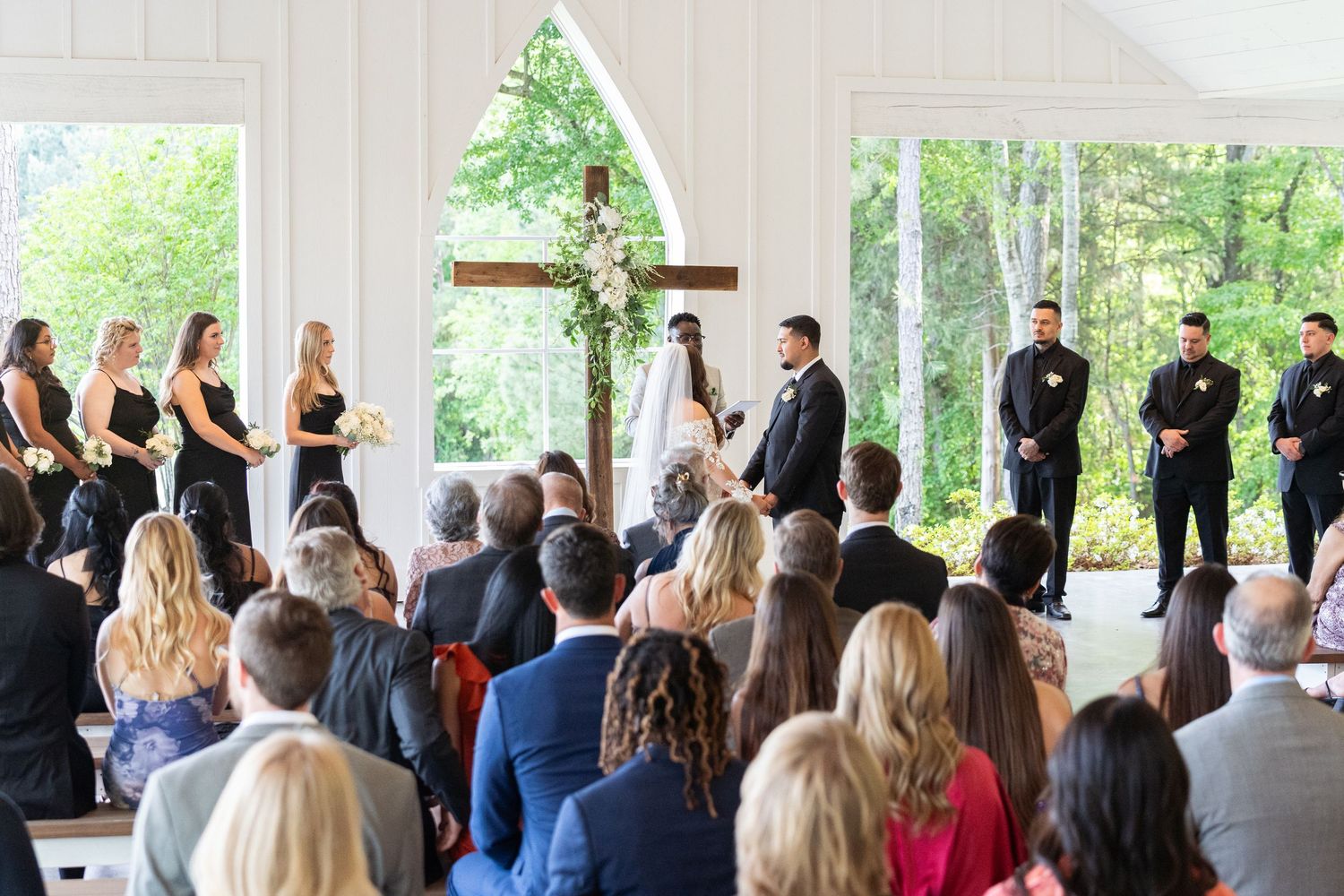 Wedding ceremony taking place under a covered outdoor pavilion with guests seated and wedding party at the altar.