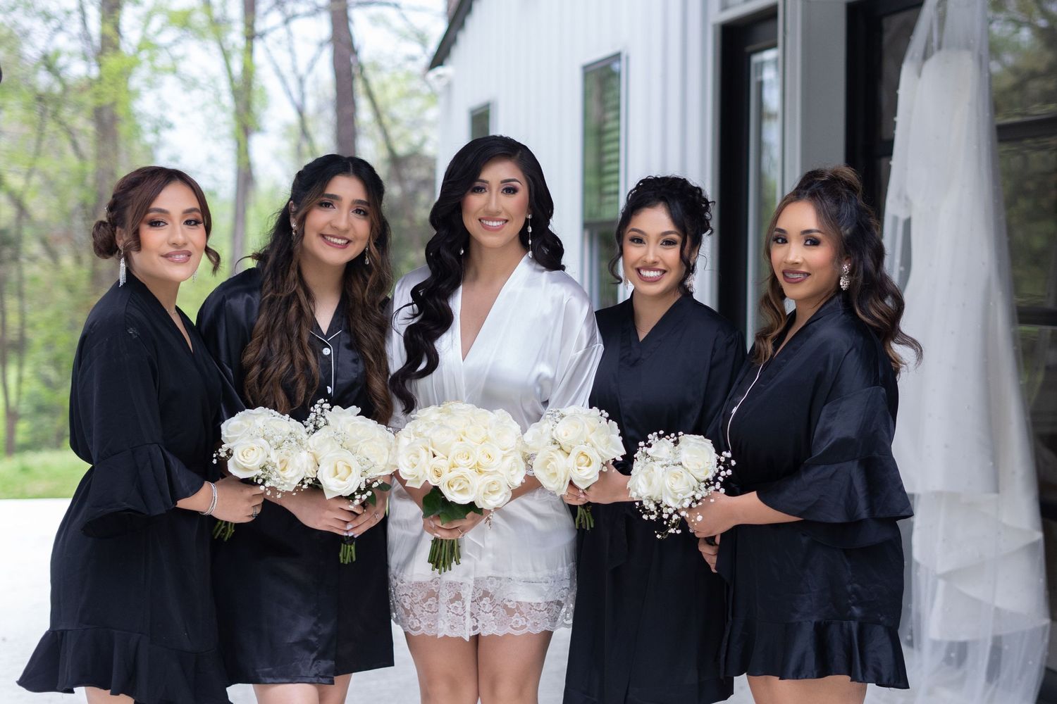 Bridesmaids in elegant black dresses pose together with white floral bouquets at an outdoor wedding venue.