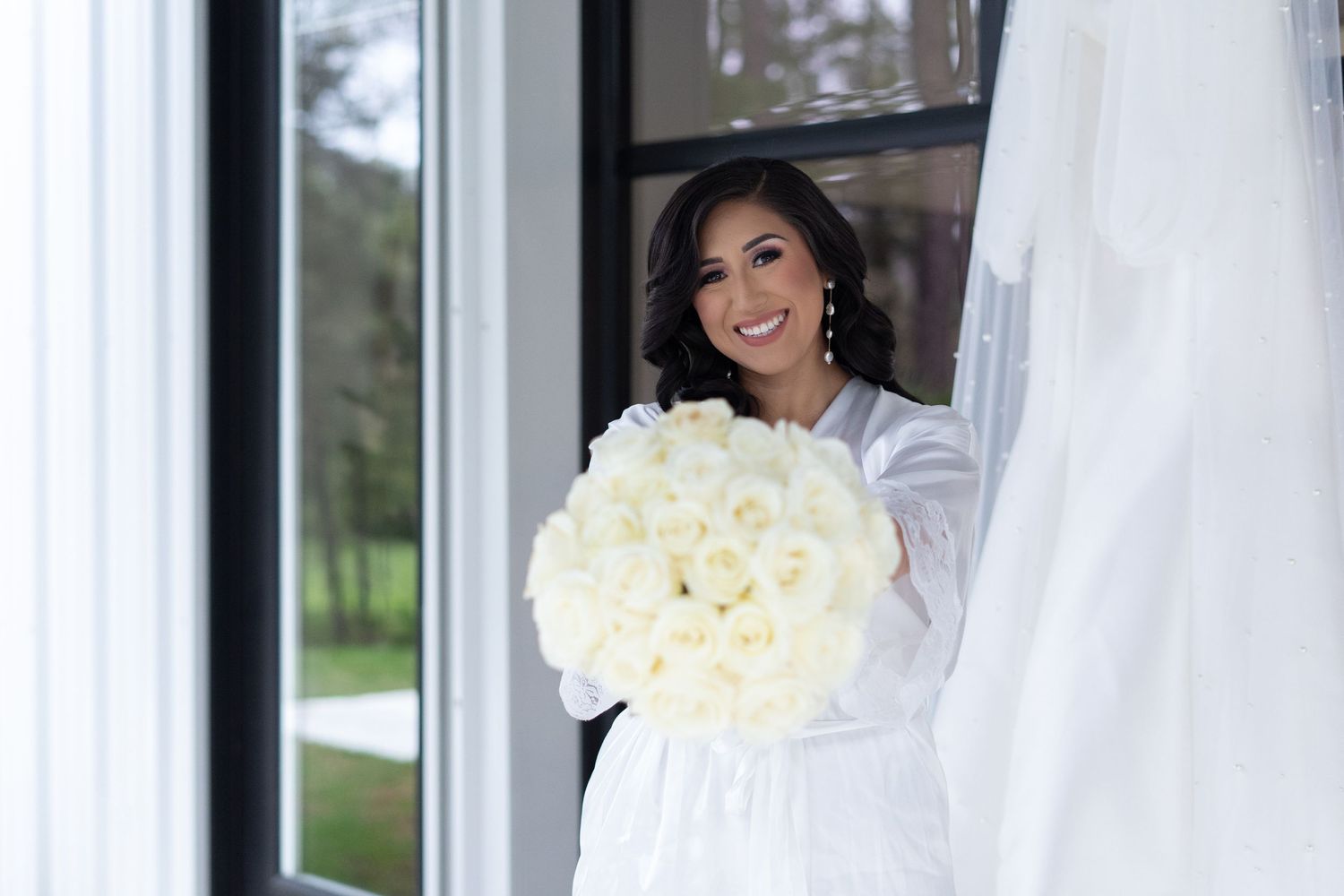 A bride in a white wedding dress holds a bouquet of cream roses while standing in front of glass windows.
