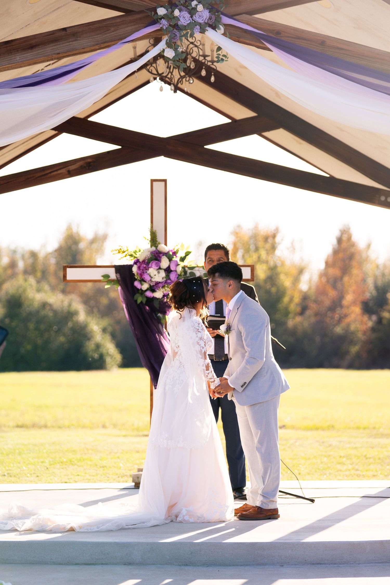 A romantic wedding ceremony scene at sunset with a couple under a wooden beam structure.