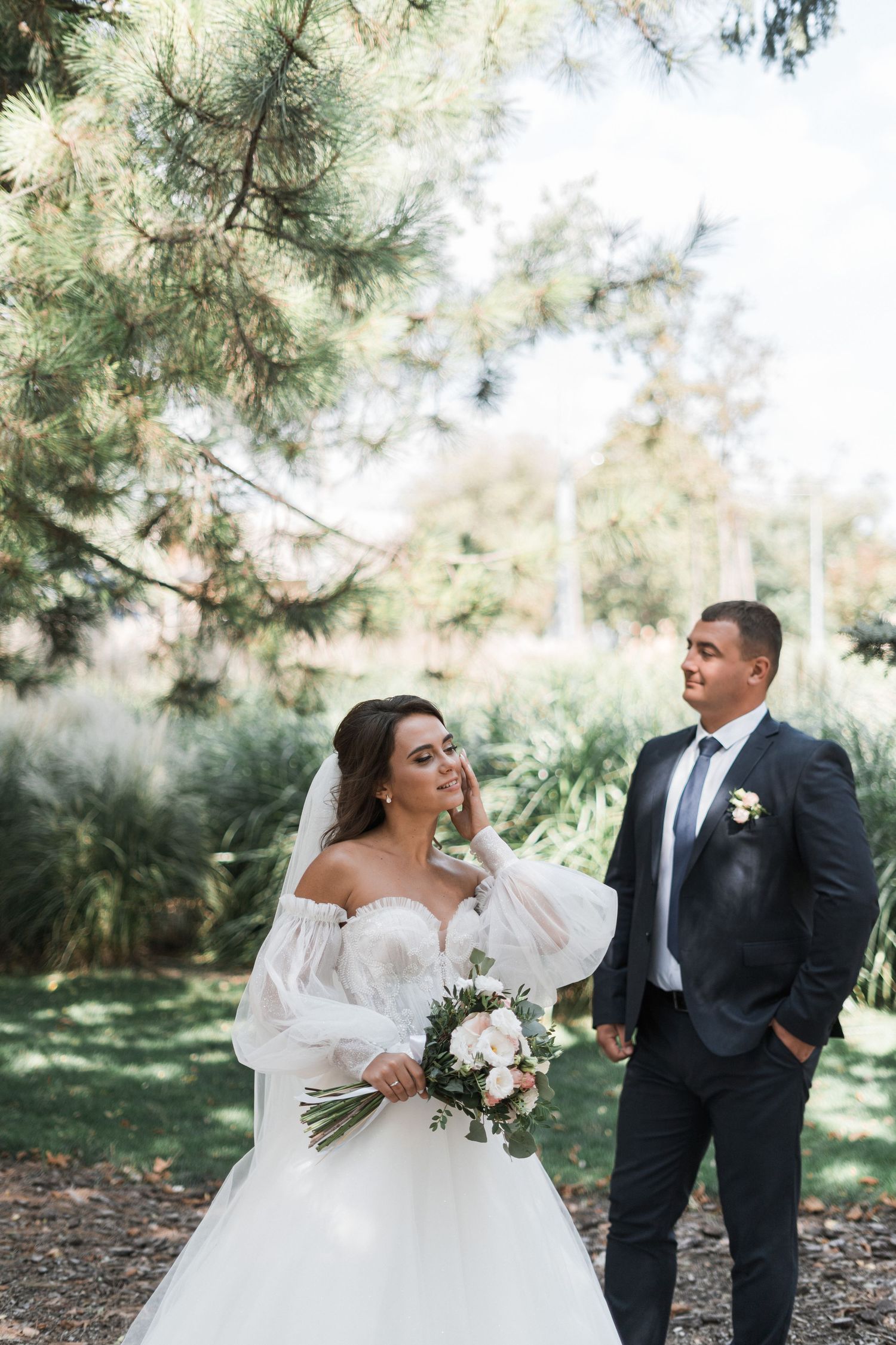 A couple in wedding attire poses outdoors under pine trees on a sunny day in a natural garden setting.
