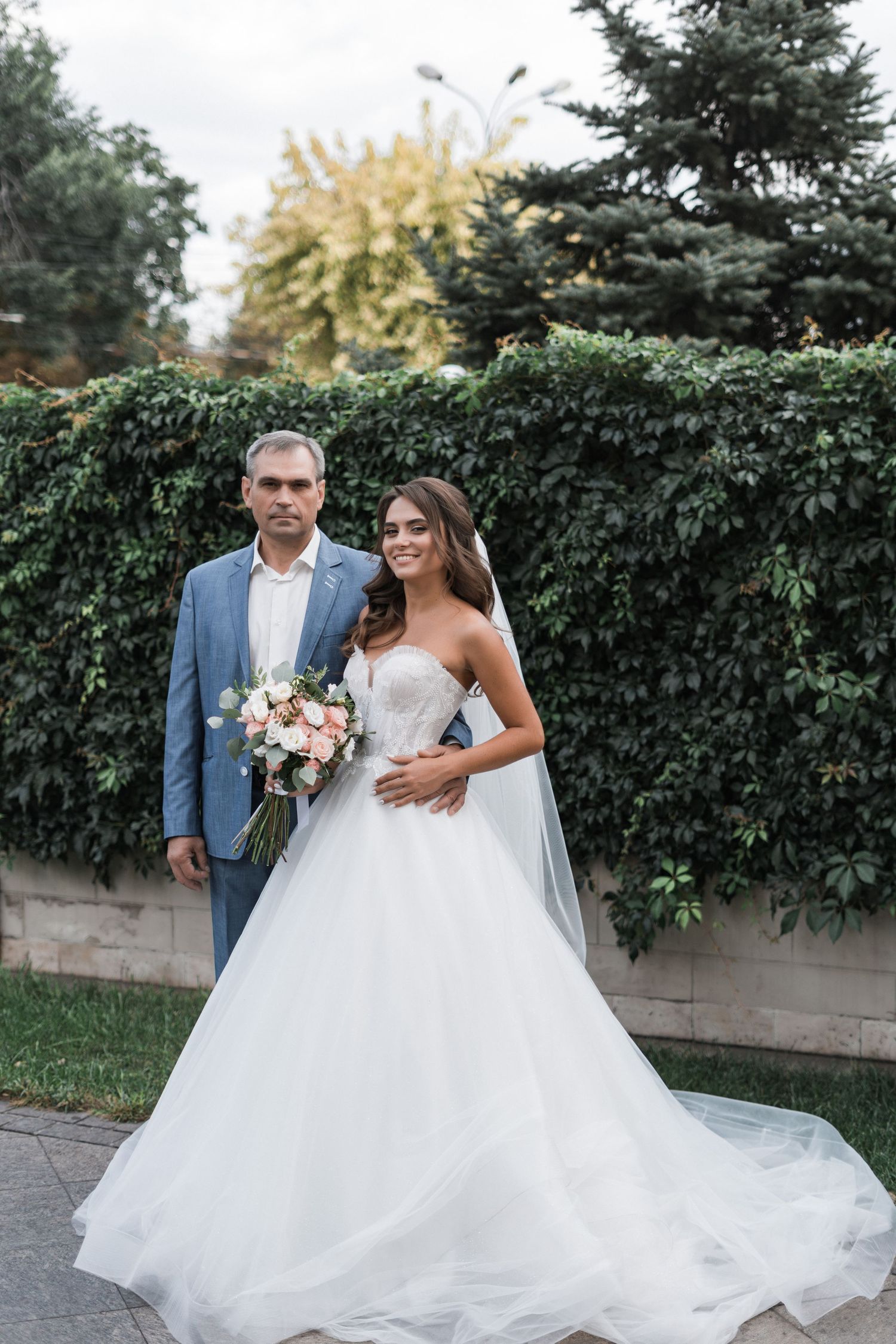 A couple in wedding attire walks together on a path beside a lush green hedge wall on their special day.