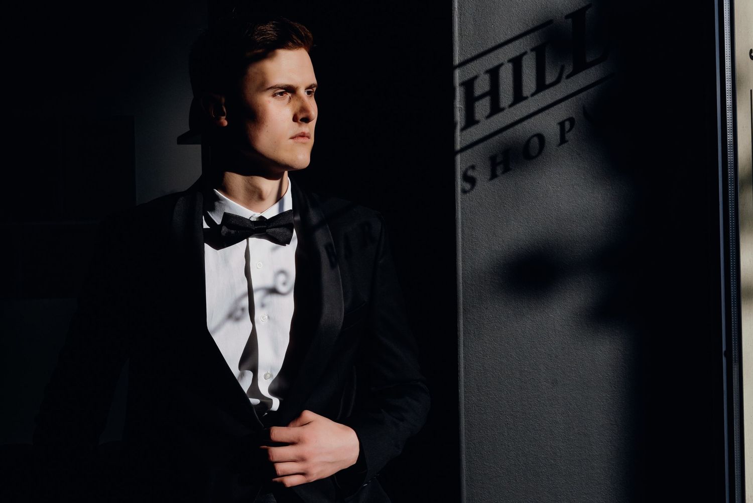 Man in formal black tuxedo and bow tie adjusts his sleeve cuff in a dramatic black and white photo.