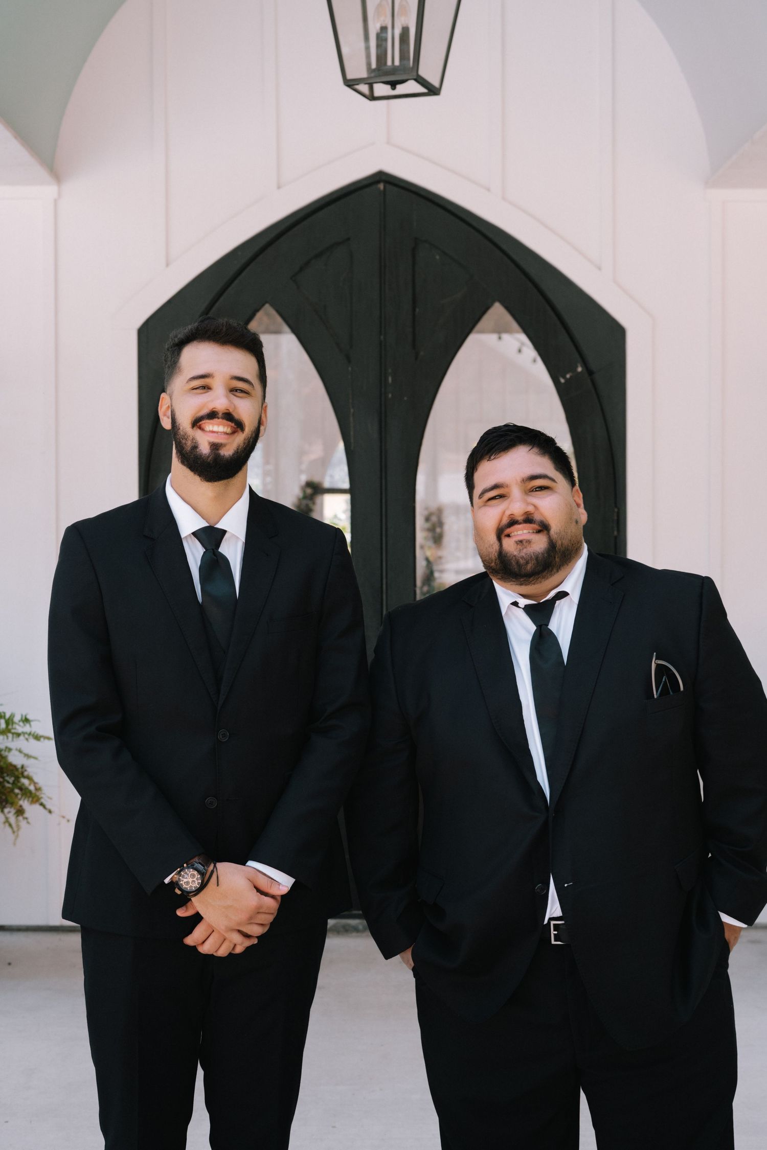 Groomsmen in black suits and ties pose together in front of a white chapel entrance.
