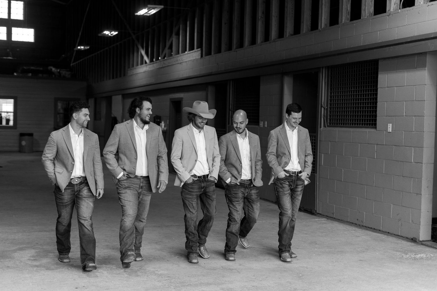 Five men in light colored blazers and jeans walk together through a dimly lit corridor.
