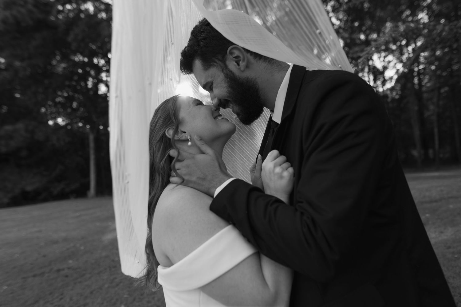 A romantic black and white wedding photo of a couple sharing a tender kiss under a veil outdoors.