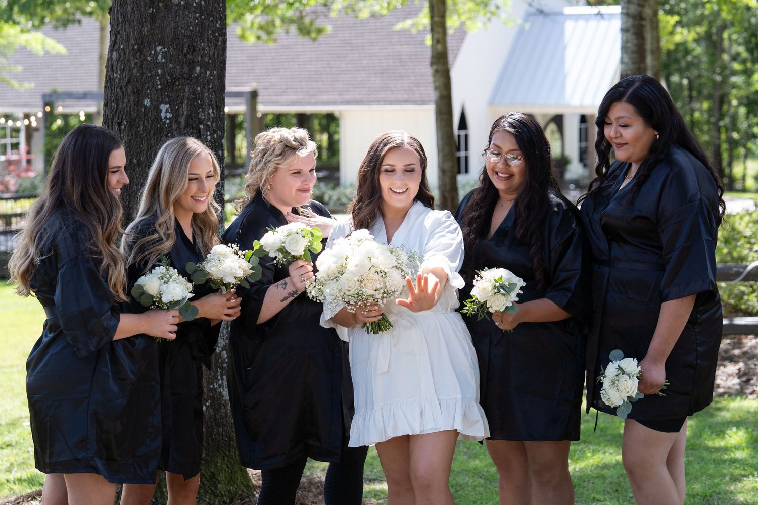 A wedding party in black and white attire poses together outdoors on a sunny day with trees in the background.