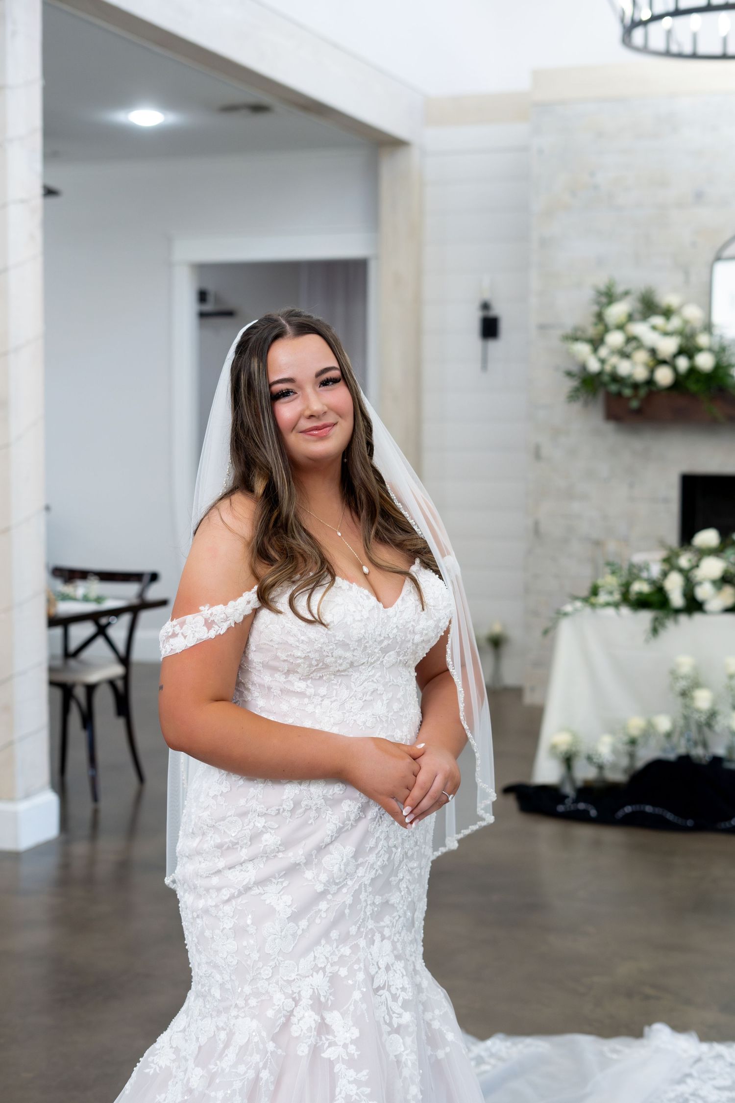 A bride in a white lace off-shoulder wedding dress poses in a bright indoor venue with white floral decorations.