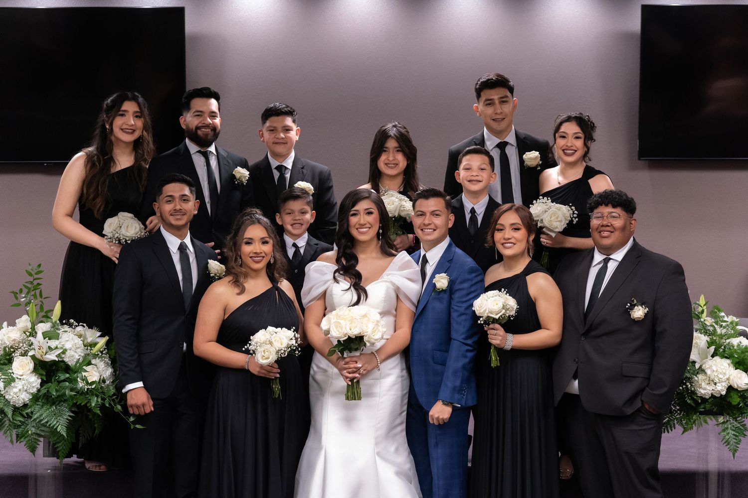 A wedding party poses together in formal attire with white floral decorations and elegant lighting in the background.