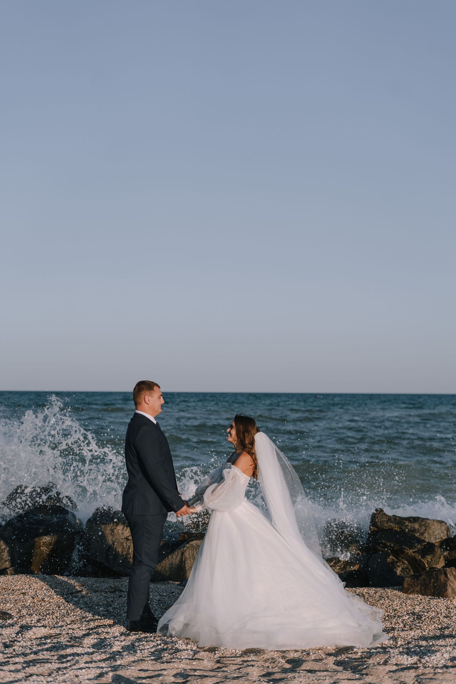 A couple in wedding attire sits on rocky beach as ocean waves crash dramatically against the shore at sunset.