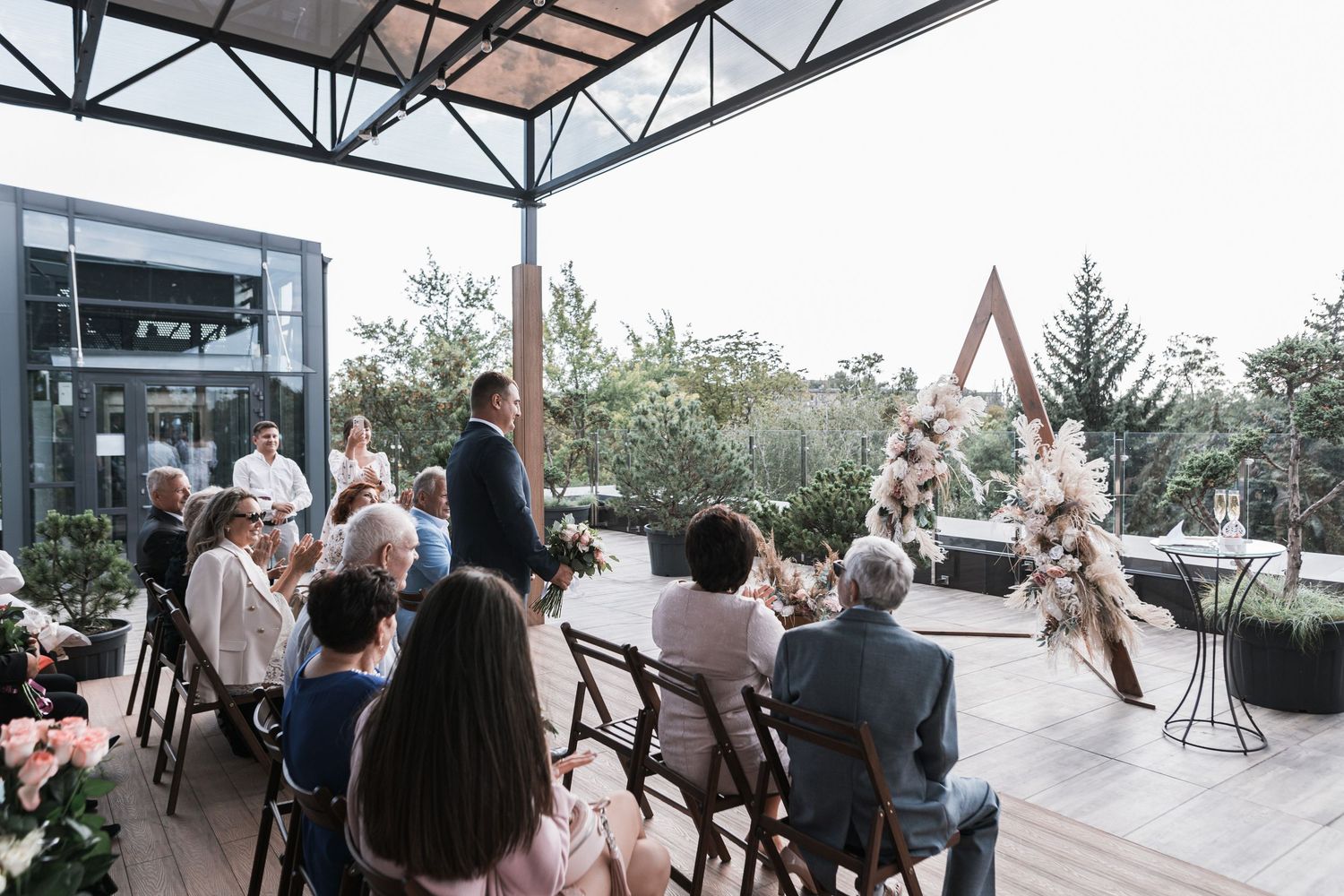 Outdoor wedding ceremony setup at a modern venue with geometric arch and floral arrangements under a covered patio.