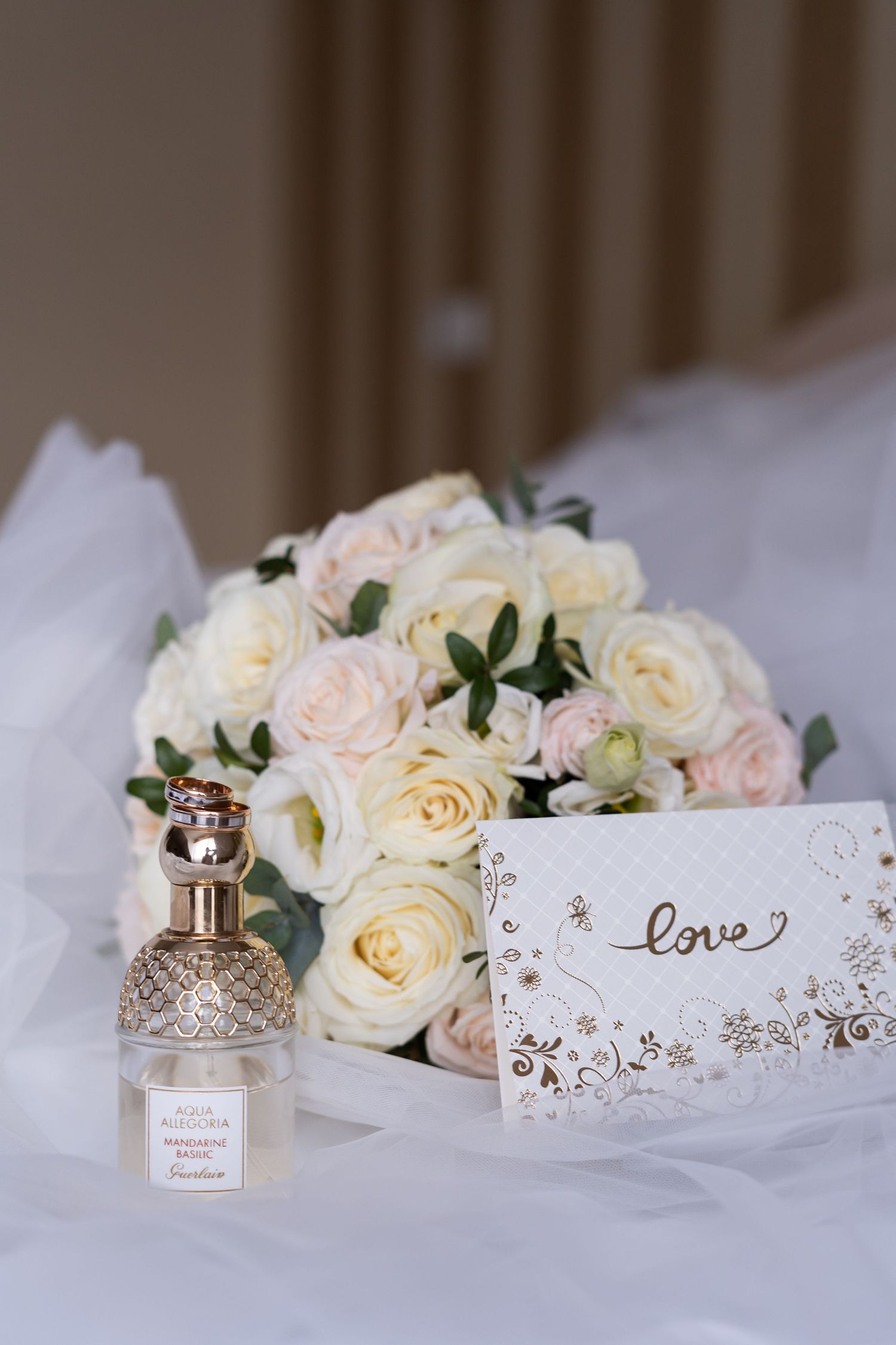 Elegant wedding details with white roses, delicate lace paper, and a crystal perfume bottle arranged on white fabric.