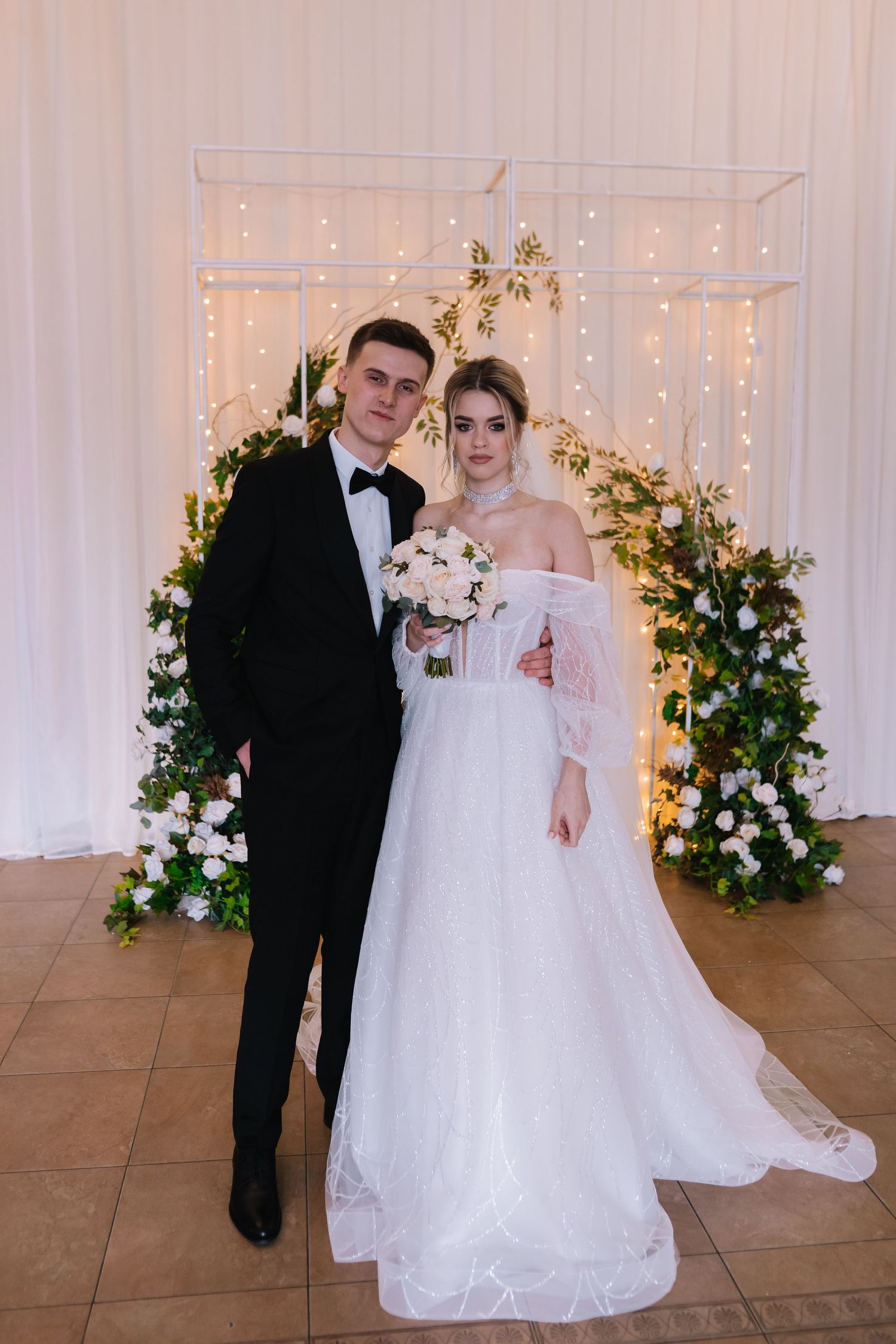 A couple poses at their wedding ceremony in front of a white backdrop with twinkling lights and floral arrangements.