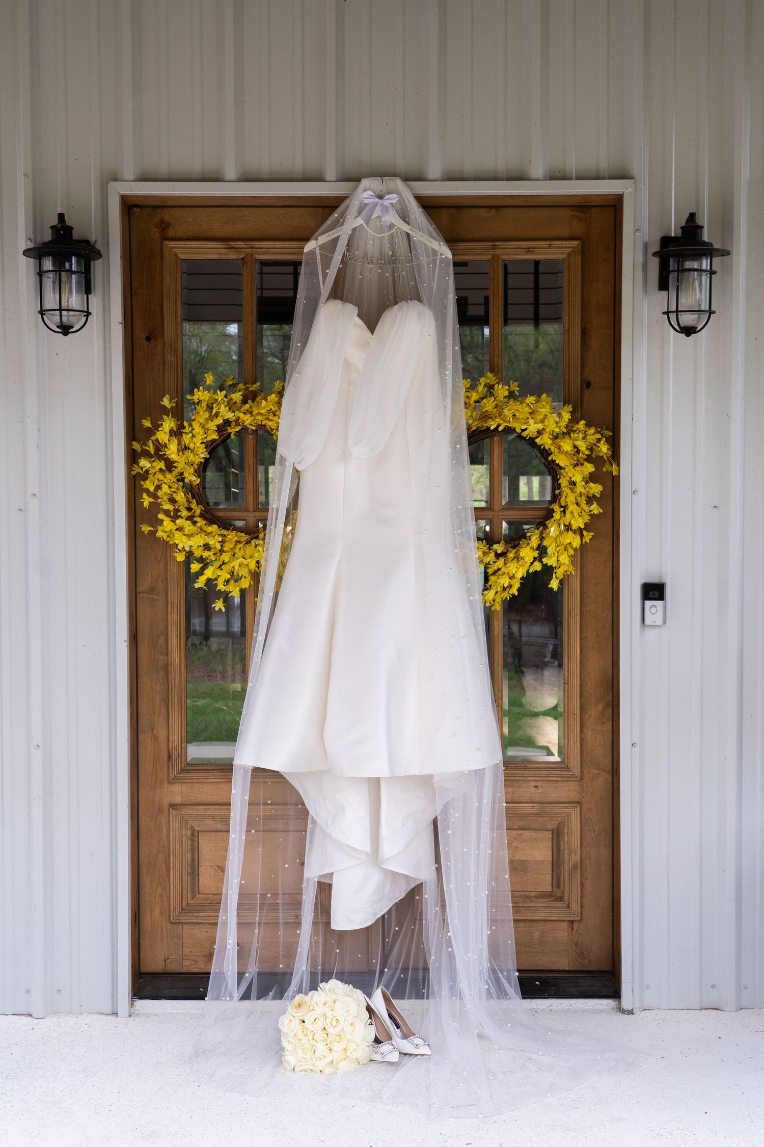 White wedding dress and veil hanging against rustic barn wall with yellow floral accents and wooden doors.