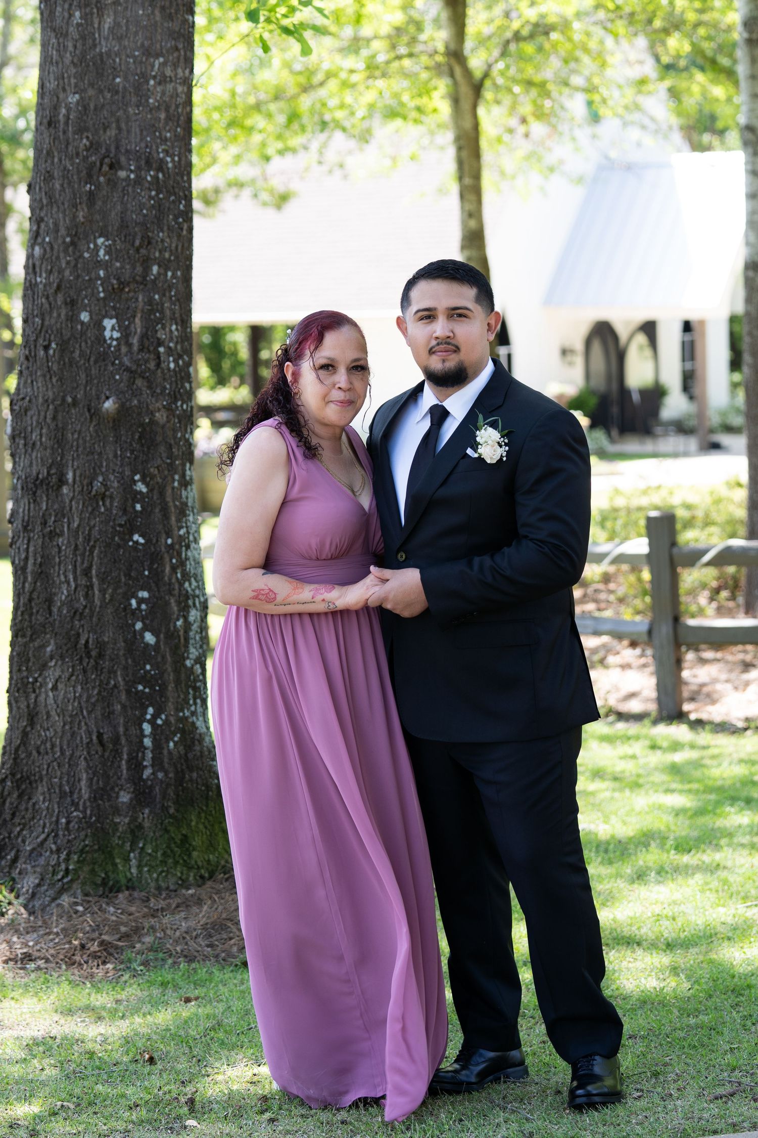 A couple in formal attire poses outdoors, with the woman wearing a lilac dress and the man in a black suit.