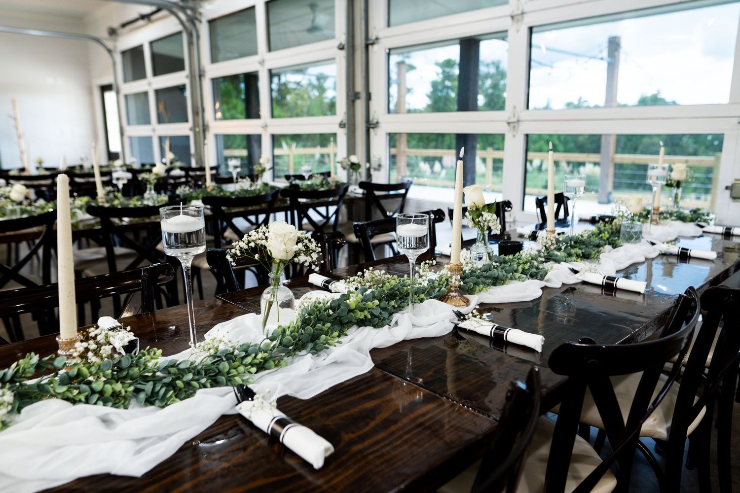 Long rustic wooden tables decorated with white runners and greenery in a bright modern reception venue with large windows.