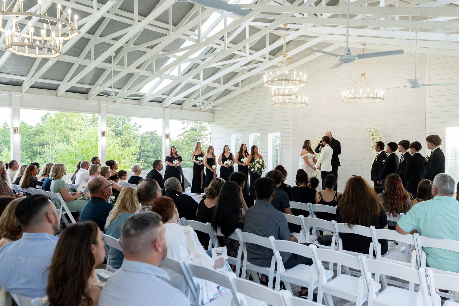 Indoor wedding ceremony in a bright white venue with exposed beams and rows of white chairs filled with guests watching the proceedings.