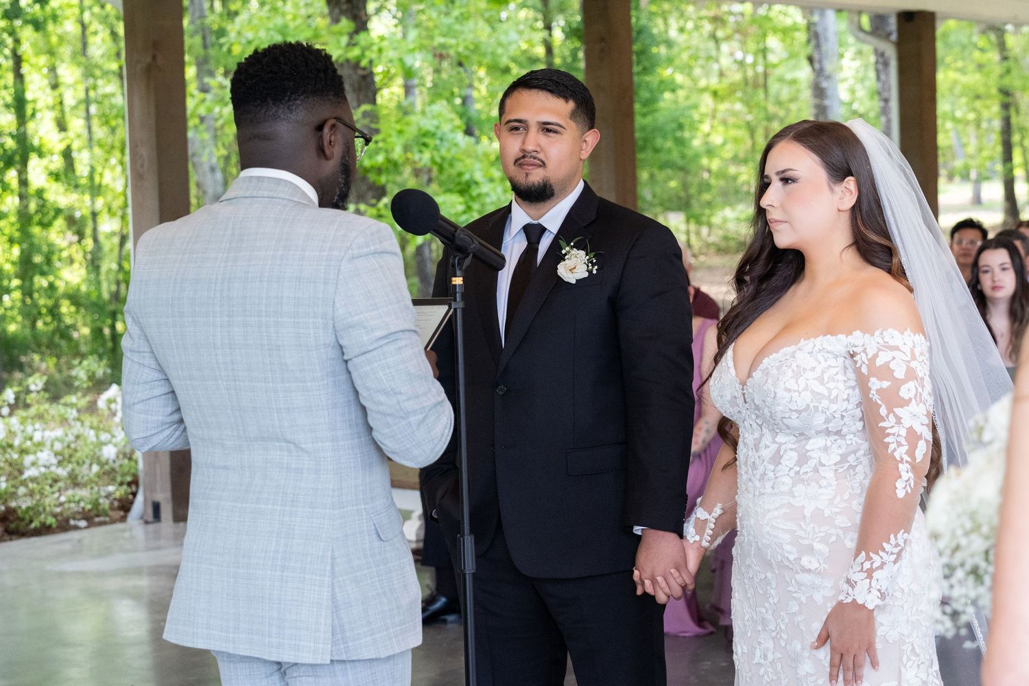 Wedding ceremony takes place outdoors under a wooden pavilion surrounded by lush green forest.