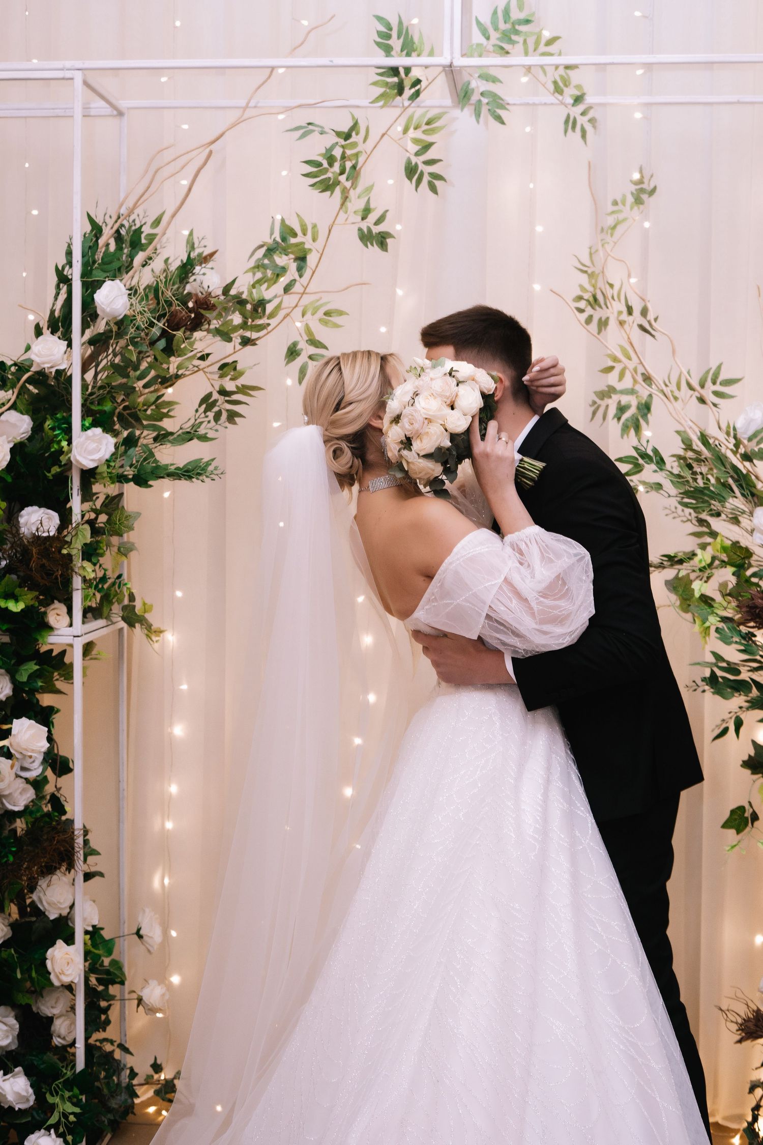 A romantic first kiss moment between newlyweds in front of a floral backdrop with twinkling lights at their wedding ceremony.