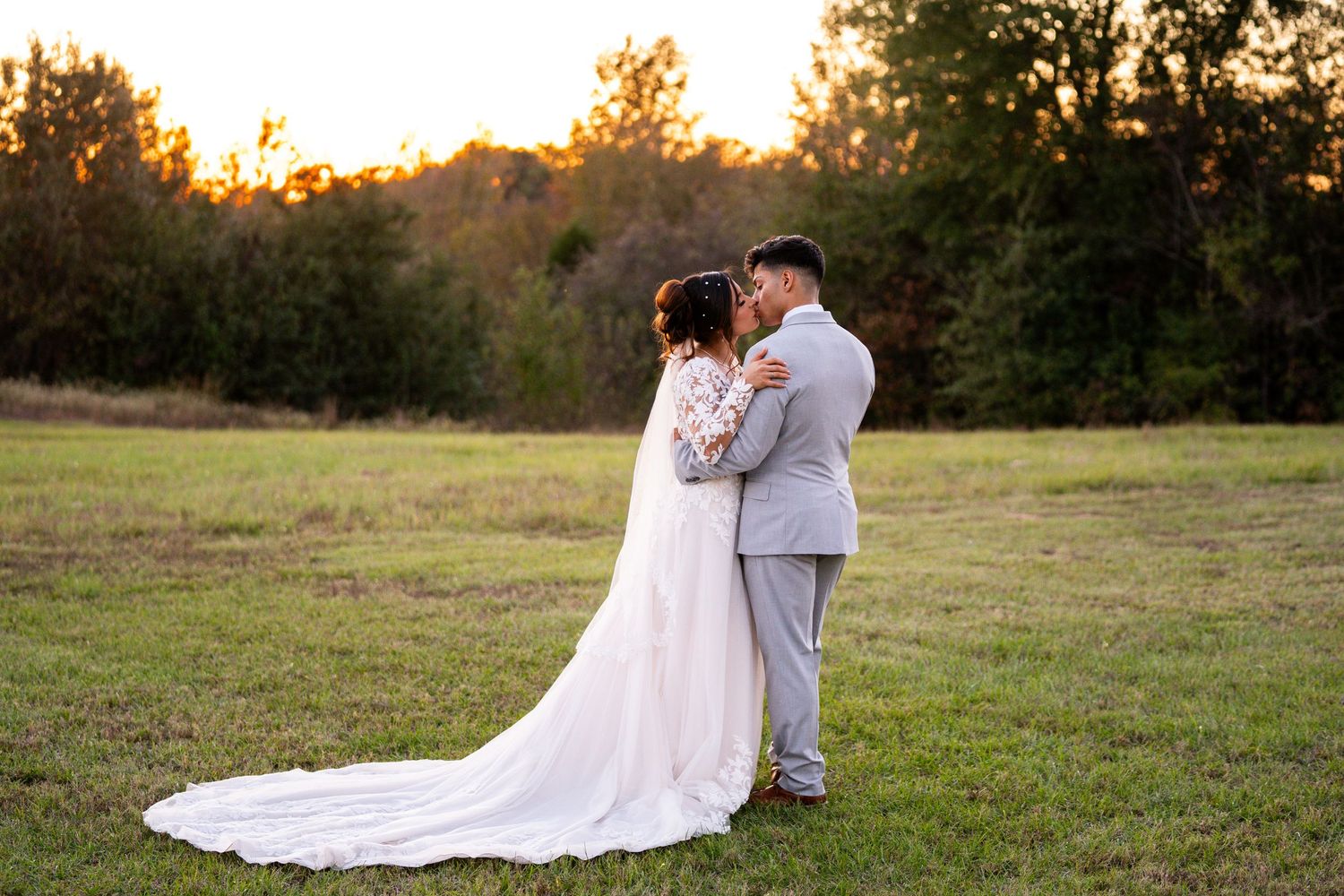 Sunset wedding portrait of couple embracing in field with long train of white dress visible.