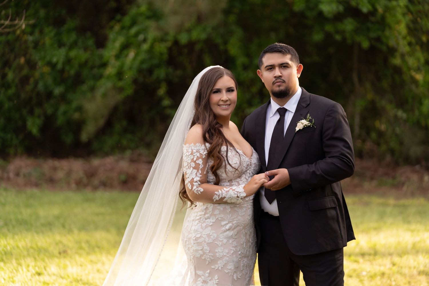 A couple in formal wedding attire poses together outdoors in a garden setting.