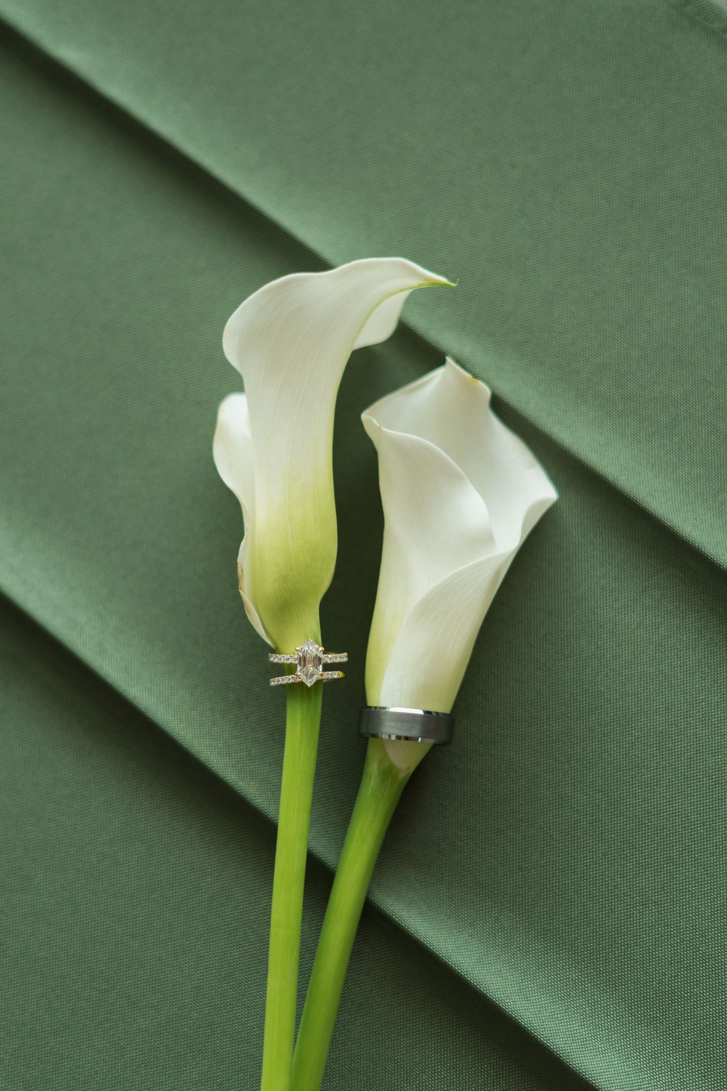 Two white calla lilies on green stems, each wearing a wedding ring, against a sage green satin background.