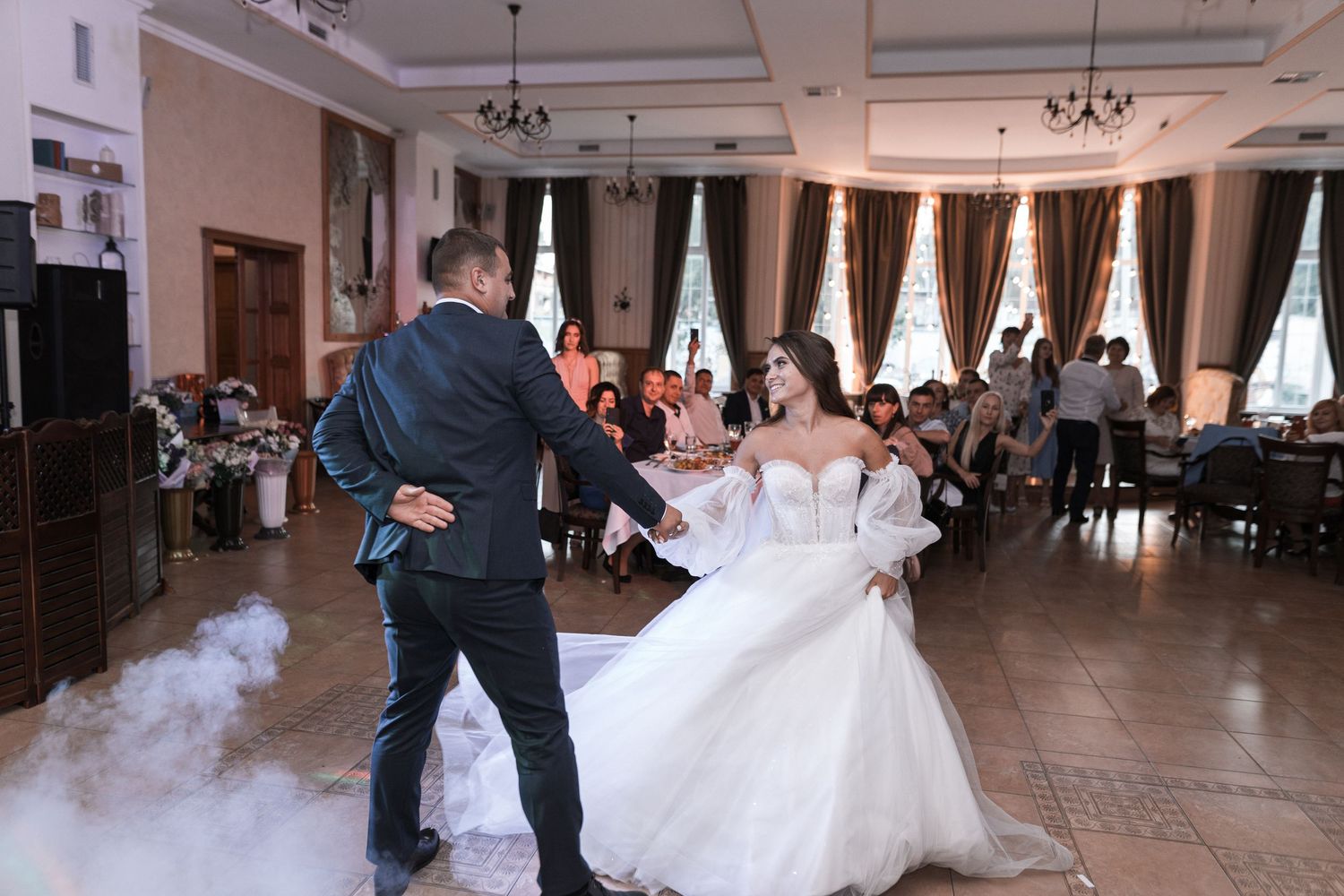 A couple performs their first dance at a wedding reception in an elegant ballroom with white decor and smoke effects.