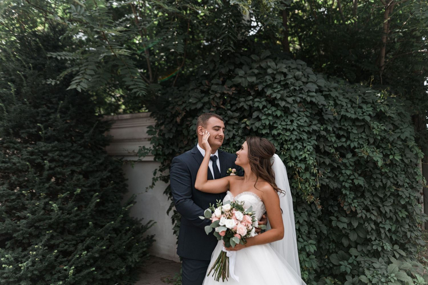 A couple in wedding attire shares a romantic moment against a lush green ivy wall backdrop.