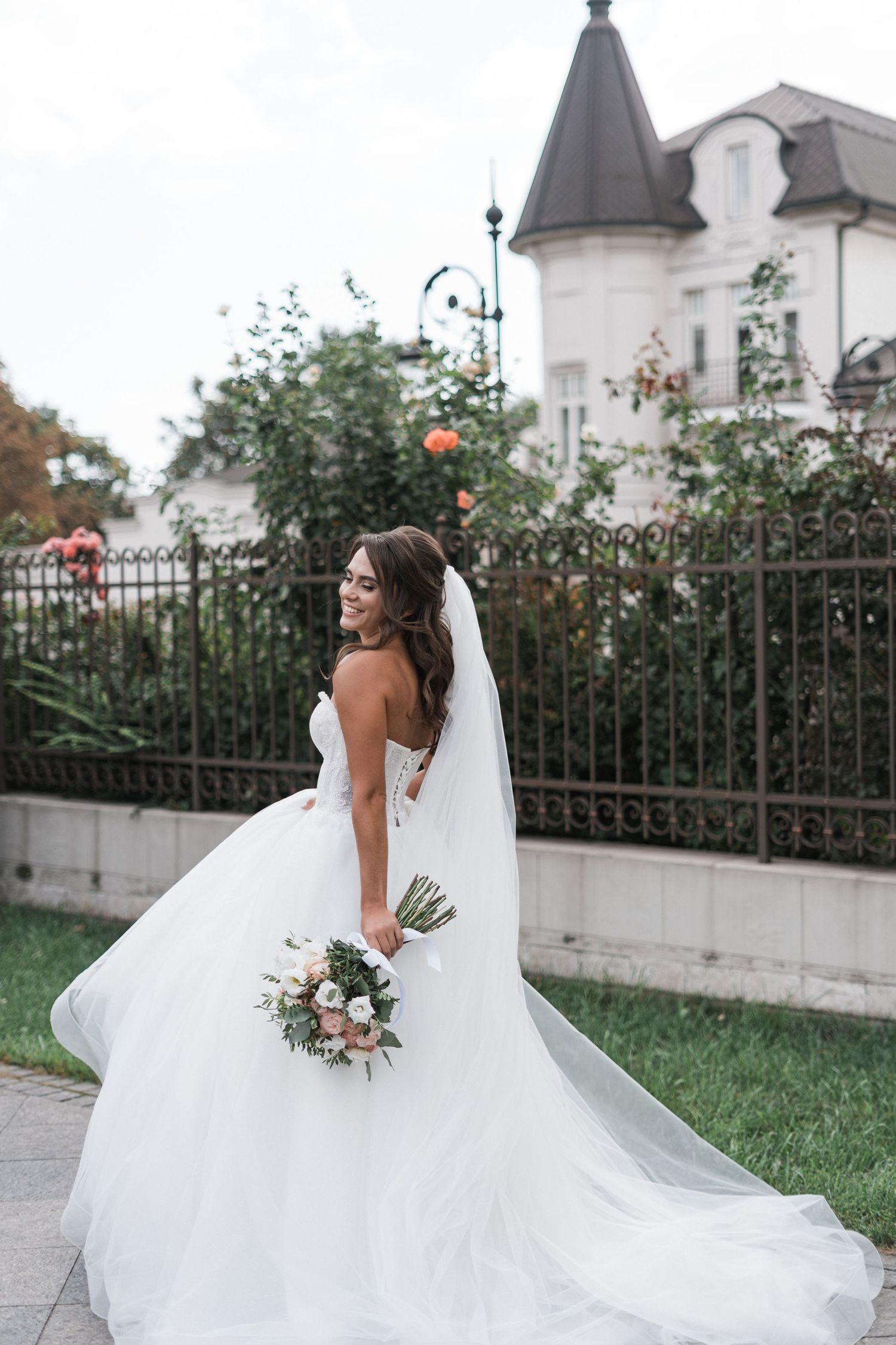 A bride in a flowing white gown with long train and veil holds a bouquet against an elegant building backdrop.