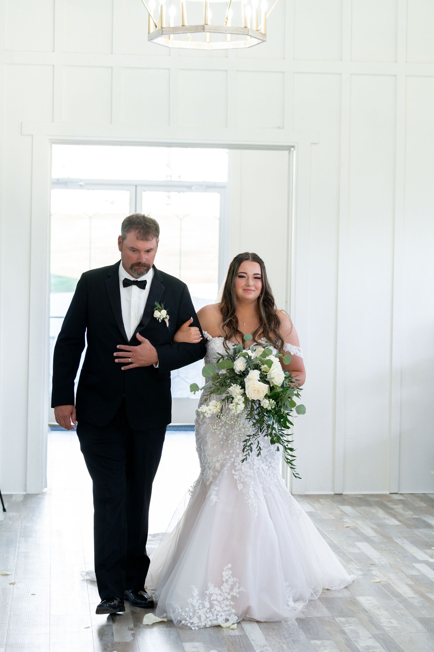 A bride and groom walk together in an elegant white room with hardwood floors and natural light streaming through windows.