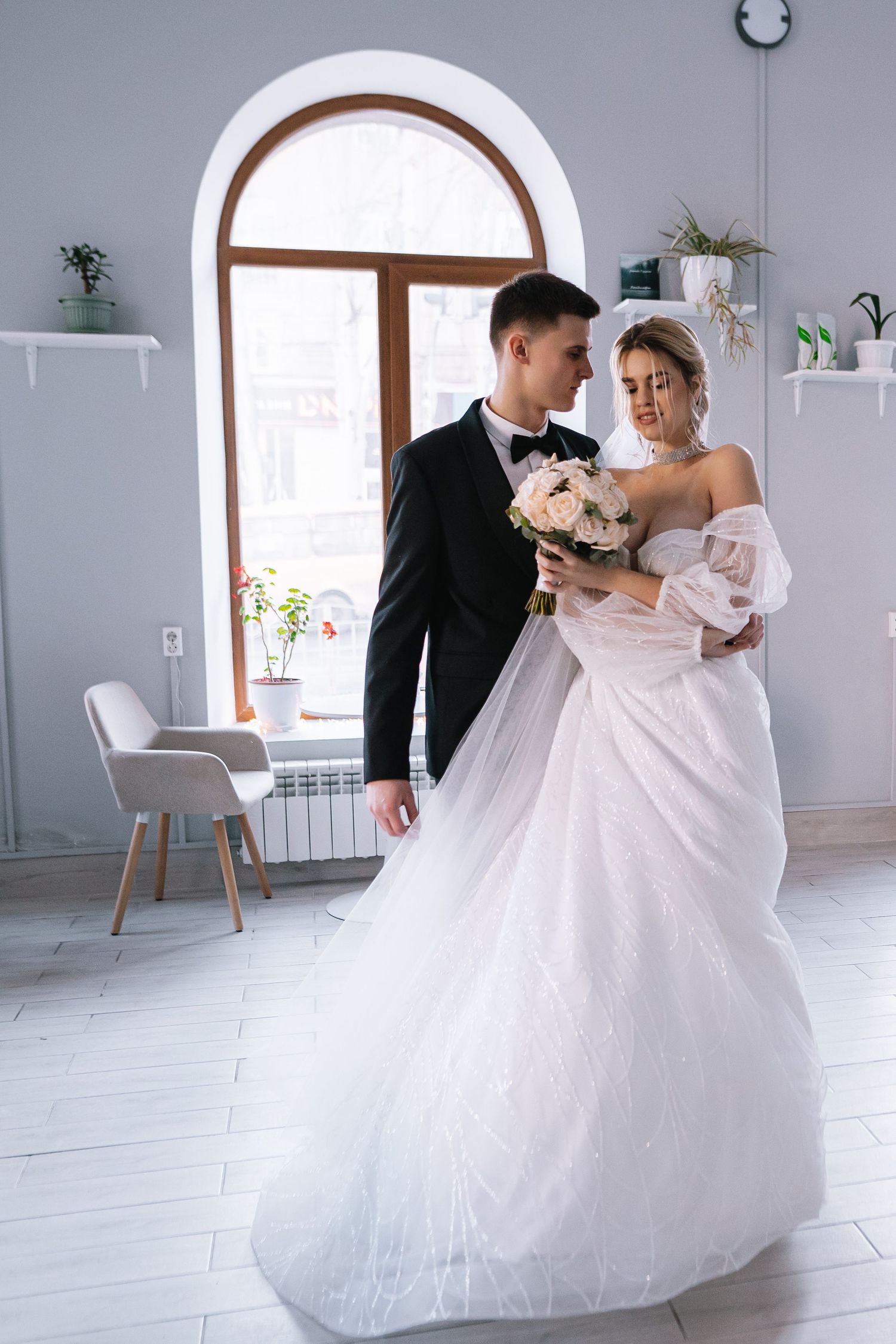 A couple in wedding attire stands in a bright white room with arched windows and modern decor.