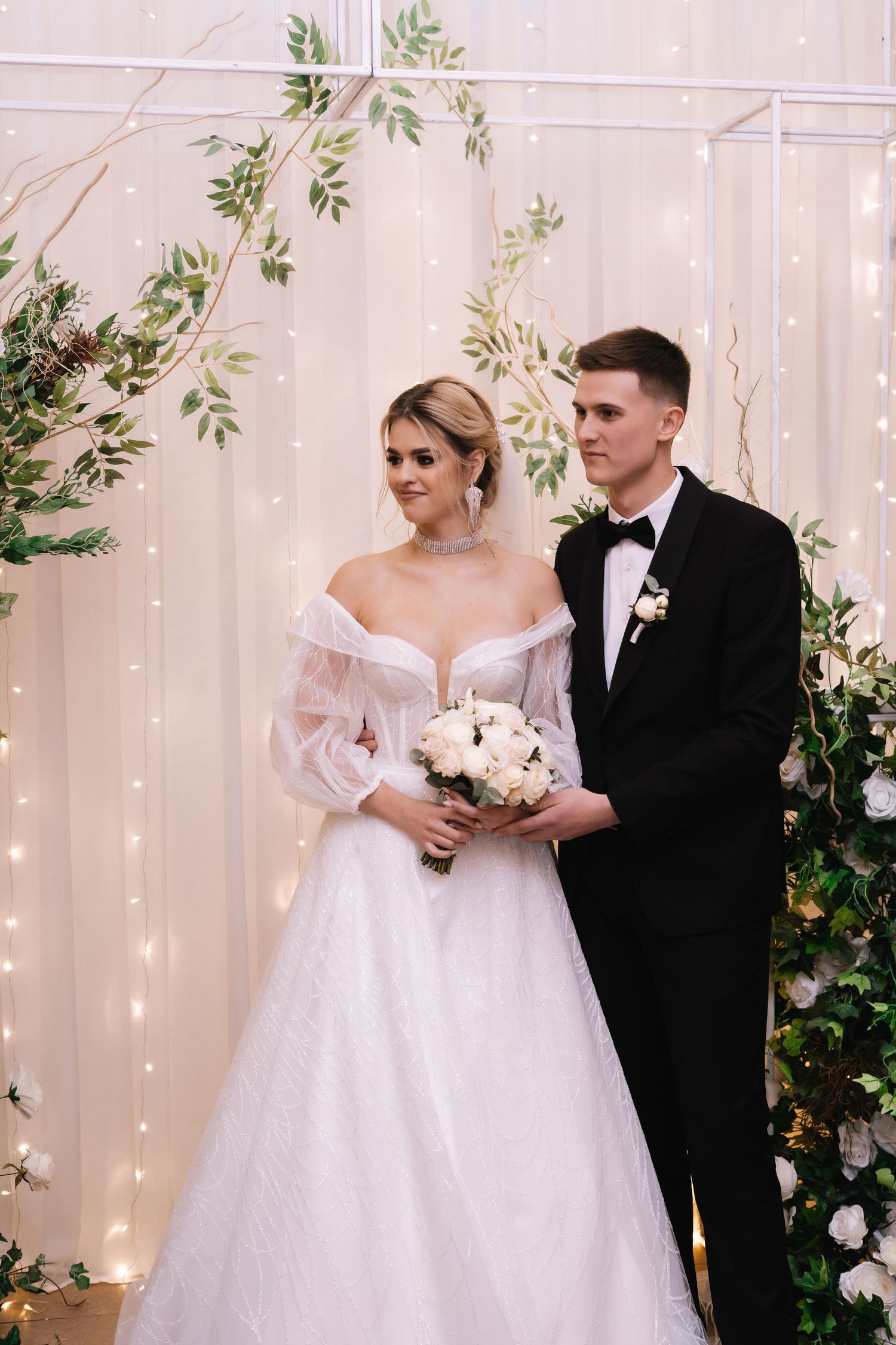A romantic wedding scene with a couple in white gown and black tuxedo posing against a fairy light backdrop with greenery.